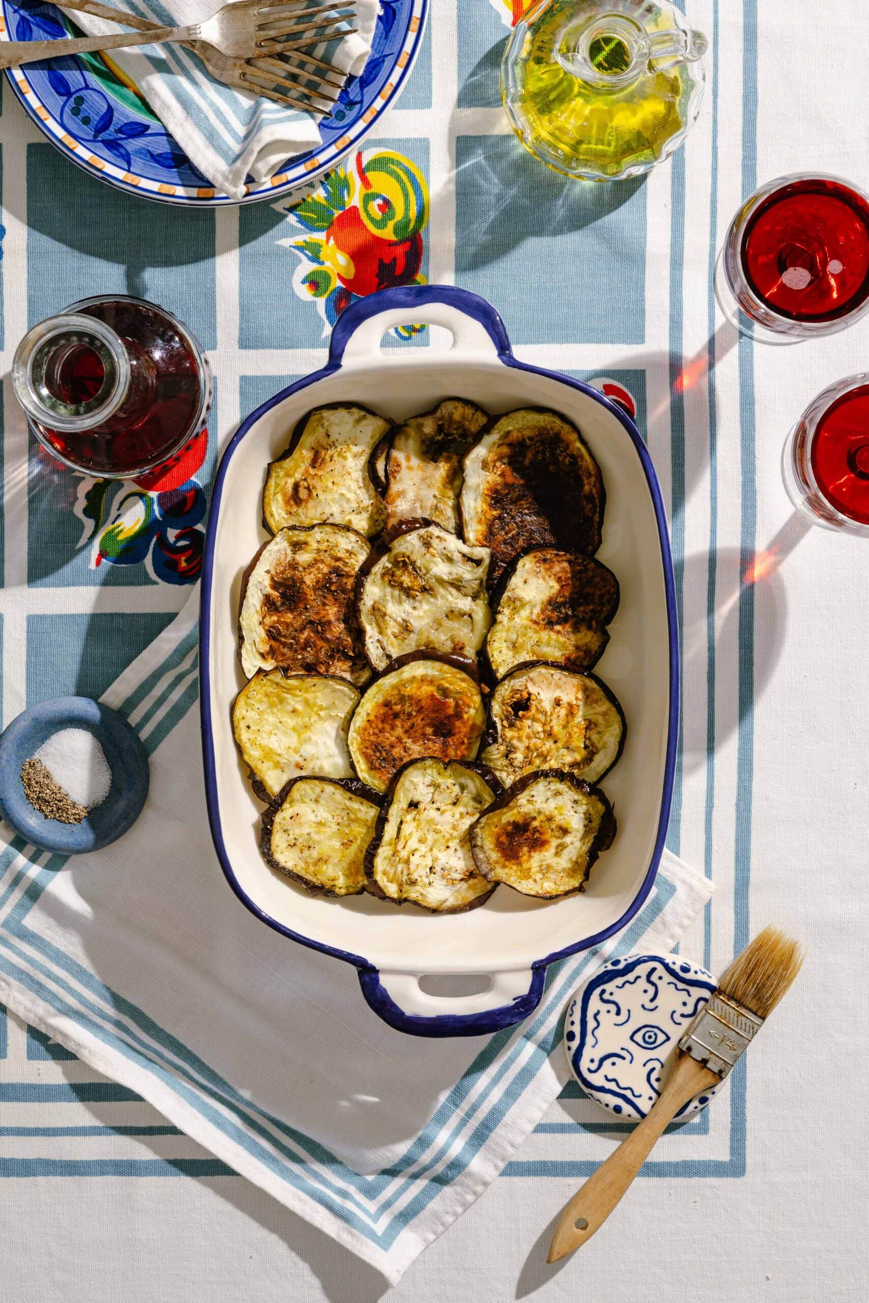 A ceramic baking dish filled with roasted eggplant slices sits on a patterned tablecloth, surrounded by wine, olive oil, a plate of silverware, a blue napkin, a small bowl of pepper, and a basting brush.