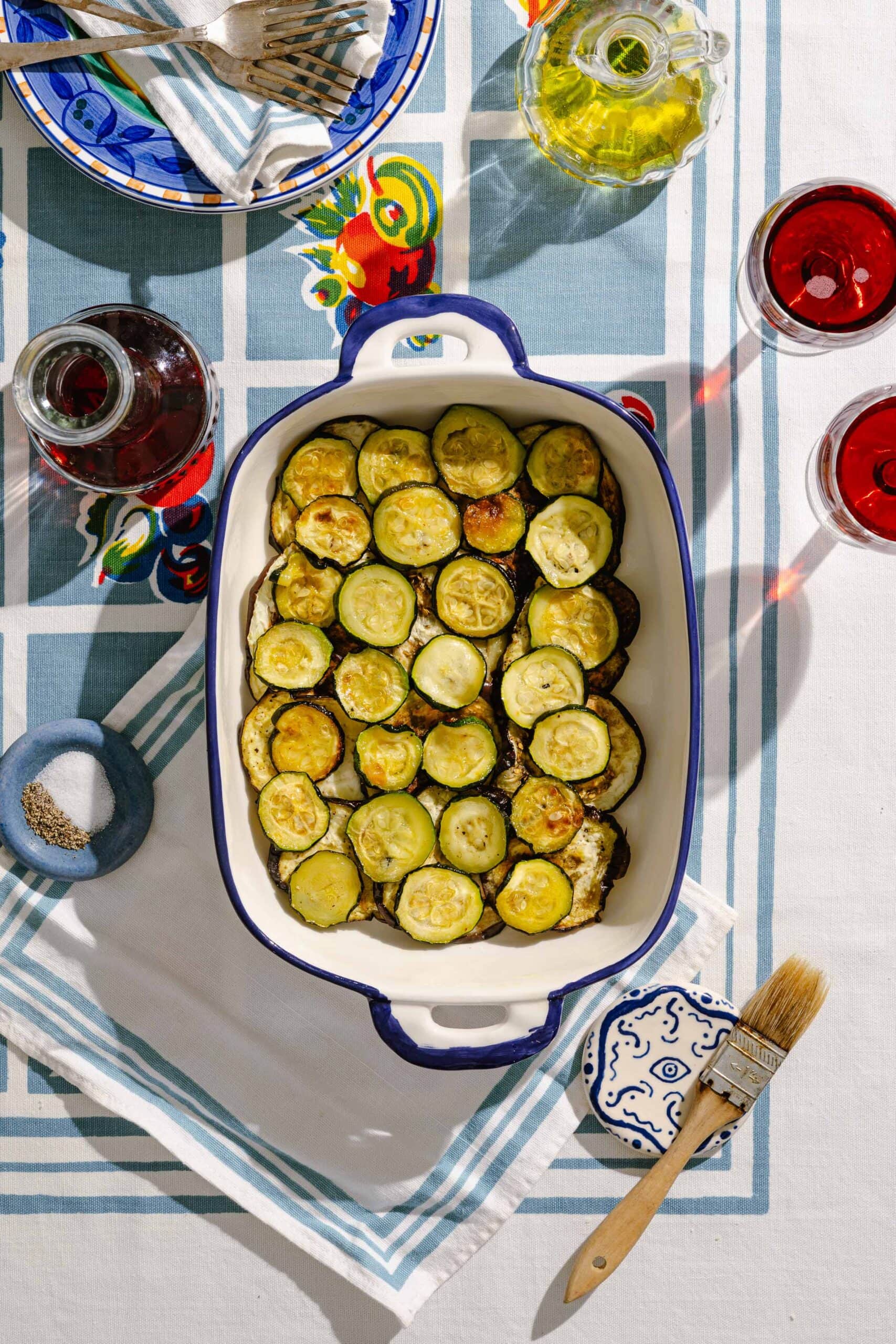A baking dish with sliced roasted zucchini sits on a table set with drinks, olive oil, a plate of utensils, a bowl of pepper, a brush, and a napkin, all arranged on a patterned tablecloth.