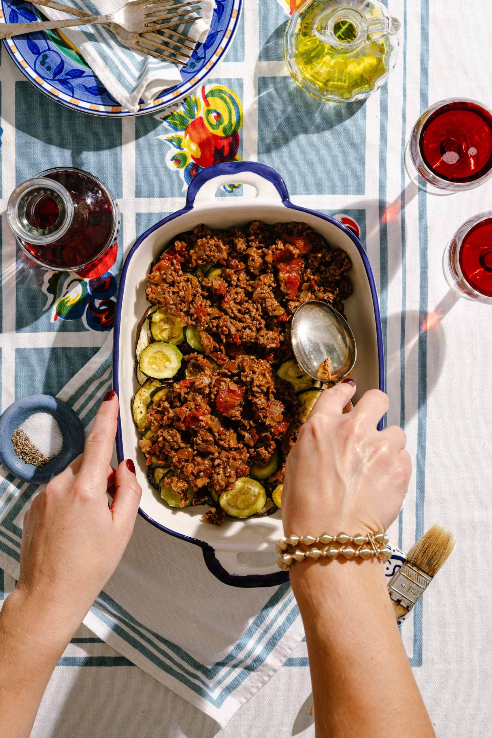 Top-down view of hands using a spoon to spread a tomato-based ground meat mixture over sliced zucchini in a blue-rimmed baking dish. The table has a blue-checkered cloth, red wine, oil, and small dishes of salt and pepper.