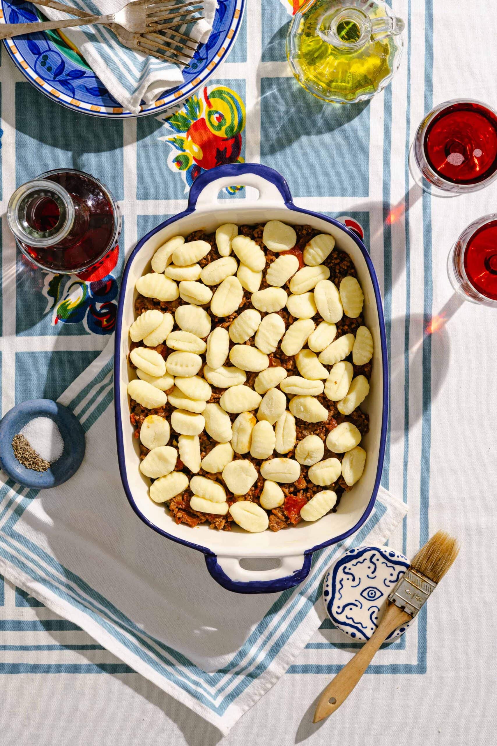 A baking dish with uncooked gnocchi over a meat sauce sits on a table set with wine, oil, utensils, and a plate, all on a colorful tablecloth.