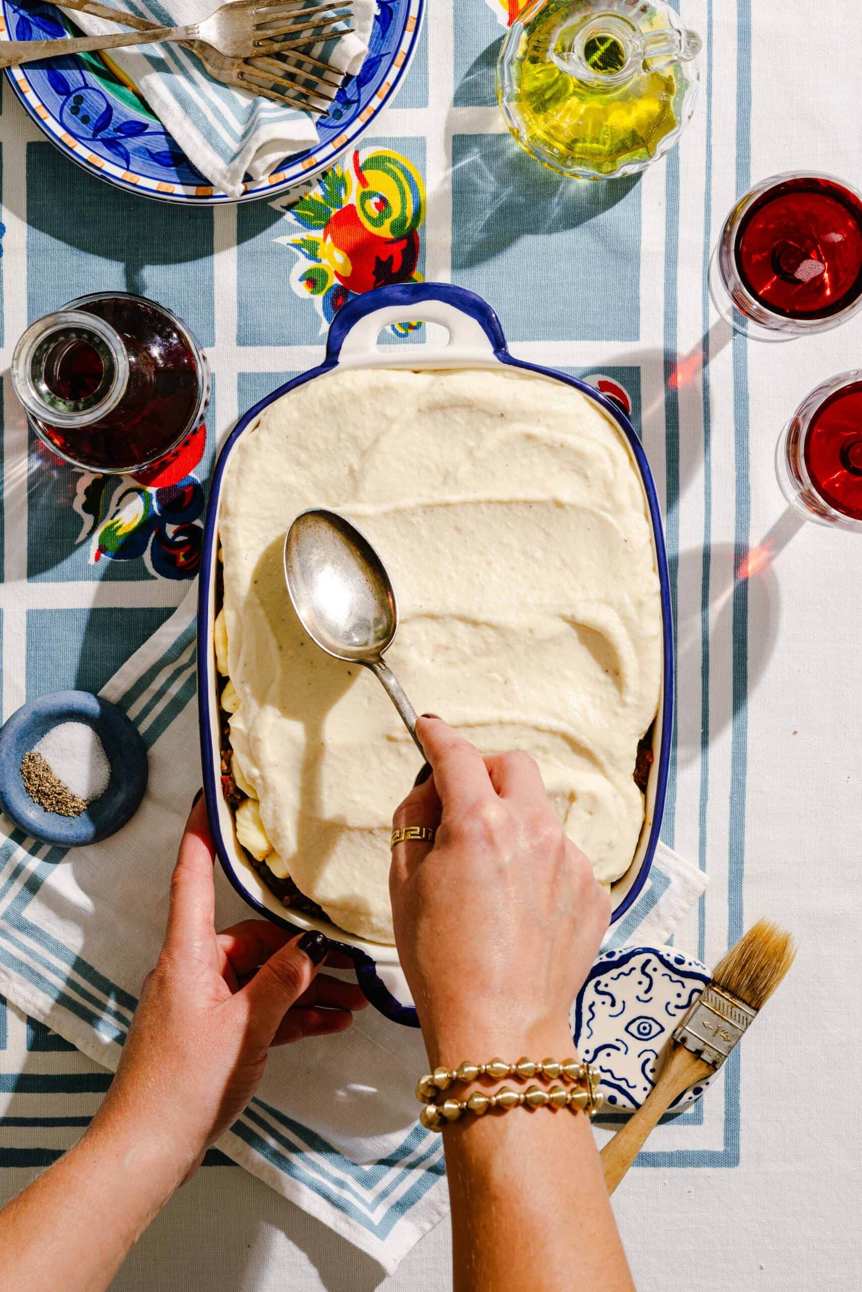 A person spreads a creamy white layer over a casserole in a blue dish on a table set with red drinks, olive oil, a bowl of forks, and a napkin.