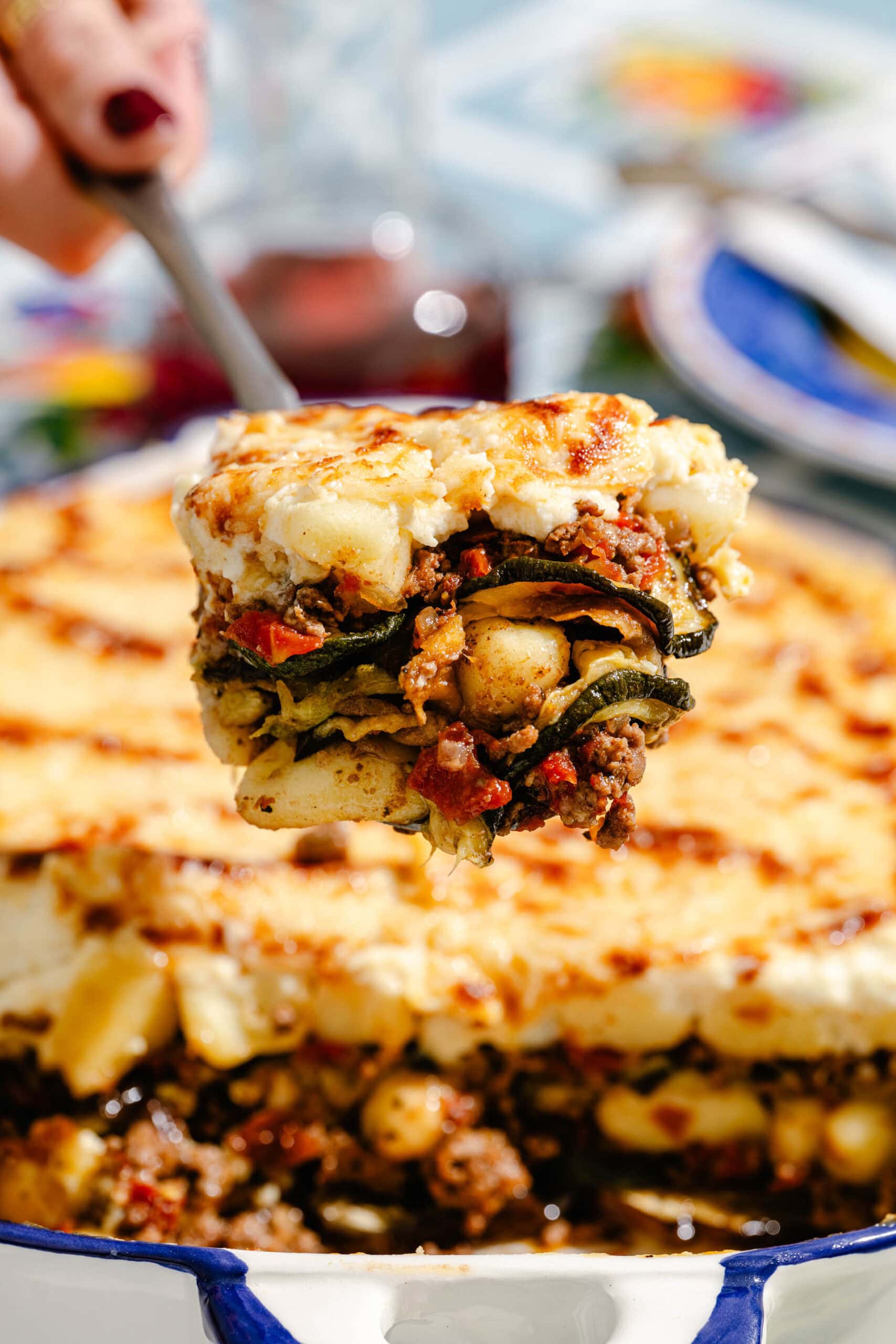 A hand lifts a serving of baked moussaka, showing layers of meat, vegetables, and creamy topping, from a casserole dish.