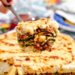 A close-up of a slice of baked lasagna with layers of meat, vegetables, and cheese being lifted from a larger dish. The background shows a colorful table setting.