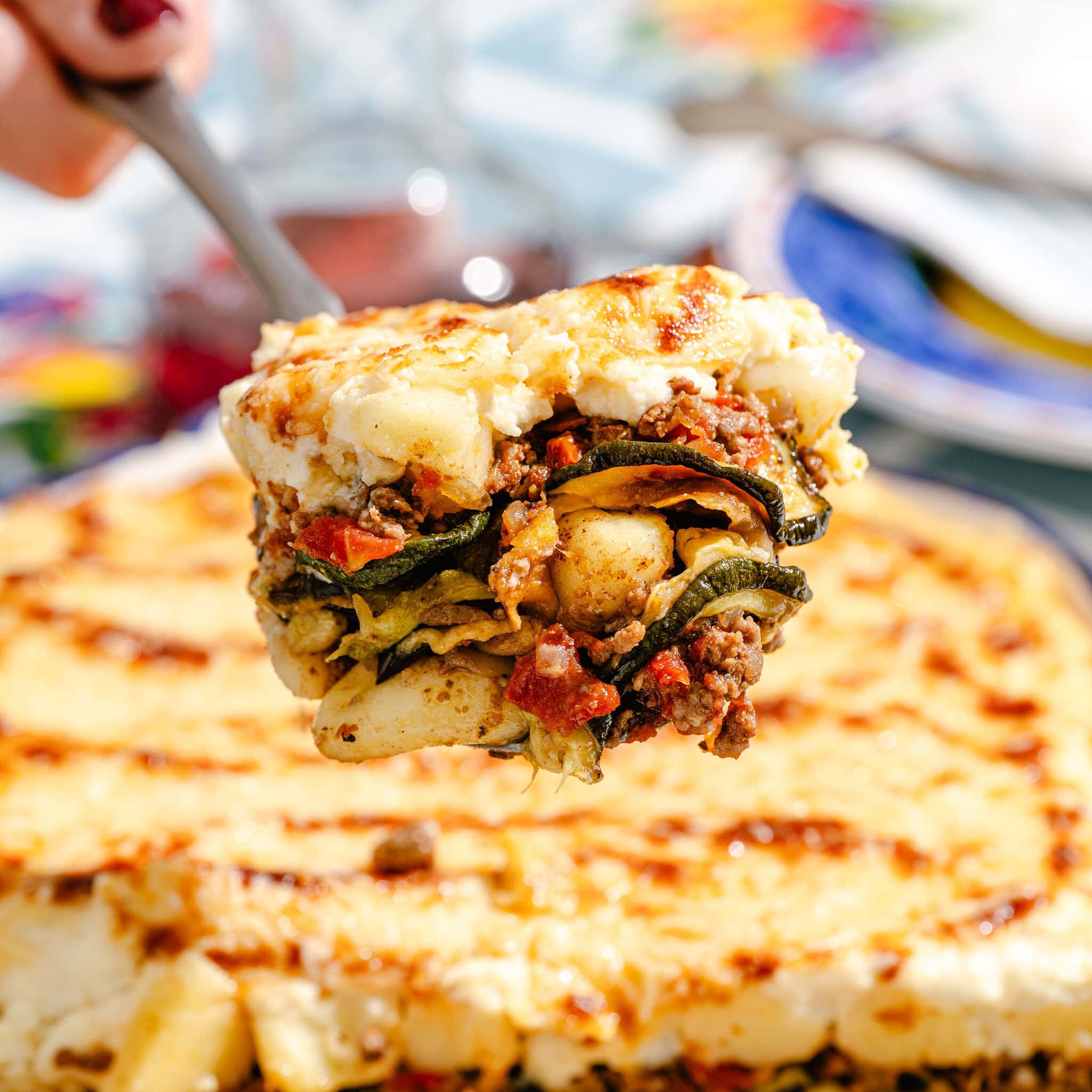 A close-up of a slice of baked lasagna with layers of meat, vegetables, and cheese being lifted from a larger dish. The background shows a colorful table setting.