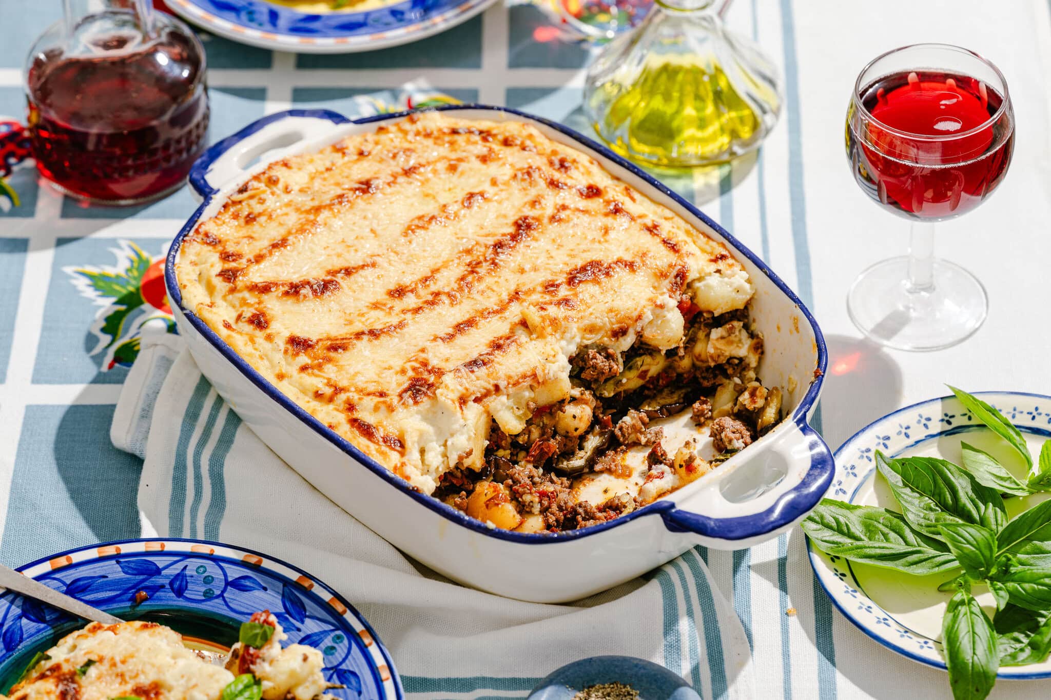 A baked casserole topped with golden cheese sits on a blue and white tablecloth, surrounded by a glass of red wine, plates, fresh basil, and bottles of oil and vinegar.
