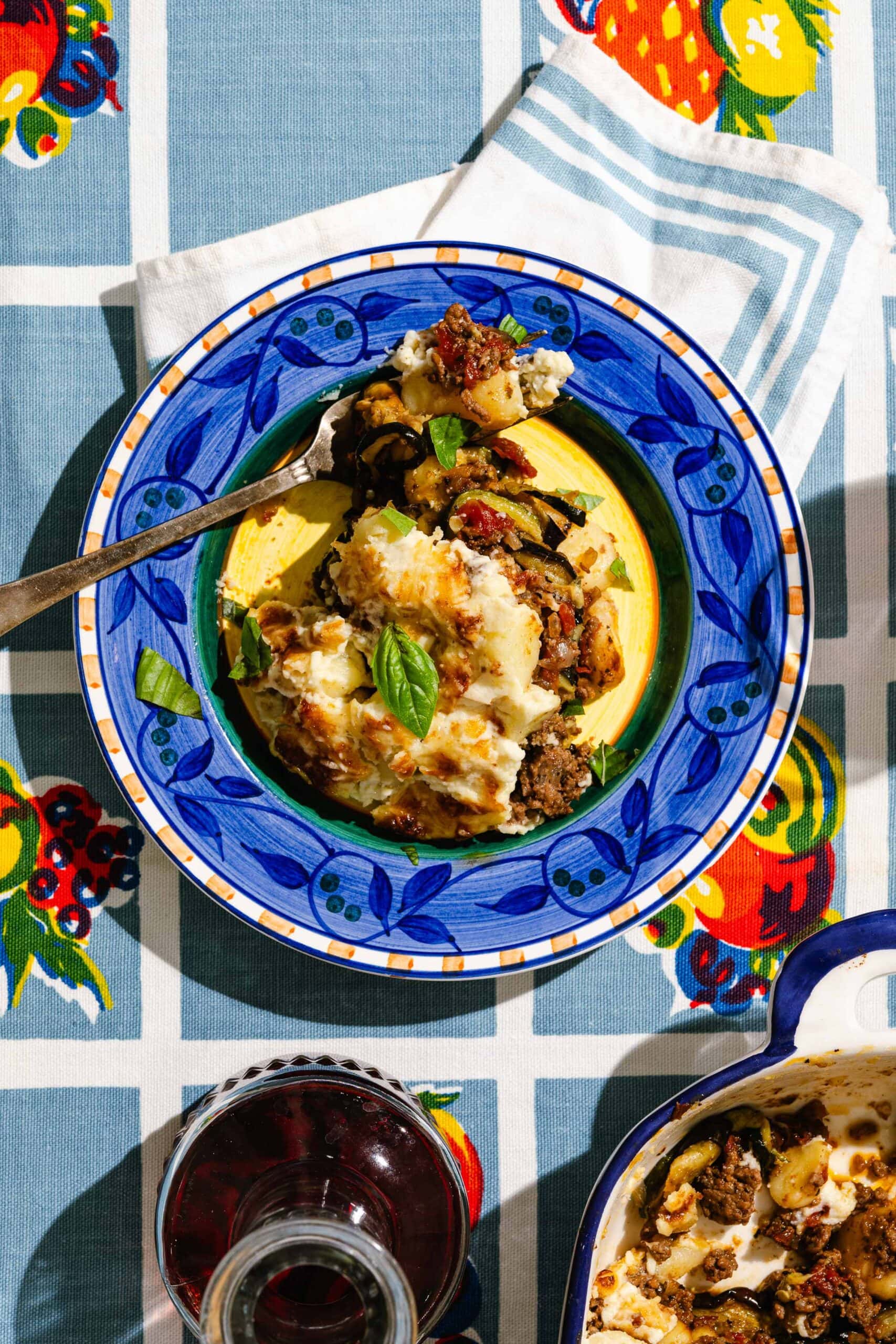 A colorful plate with a serving of baked casserole, garnished with basil, sits on a patterned tablecloth next to a glass of red wine and a napkin. A fork rests on the plate.