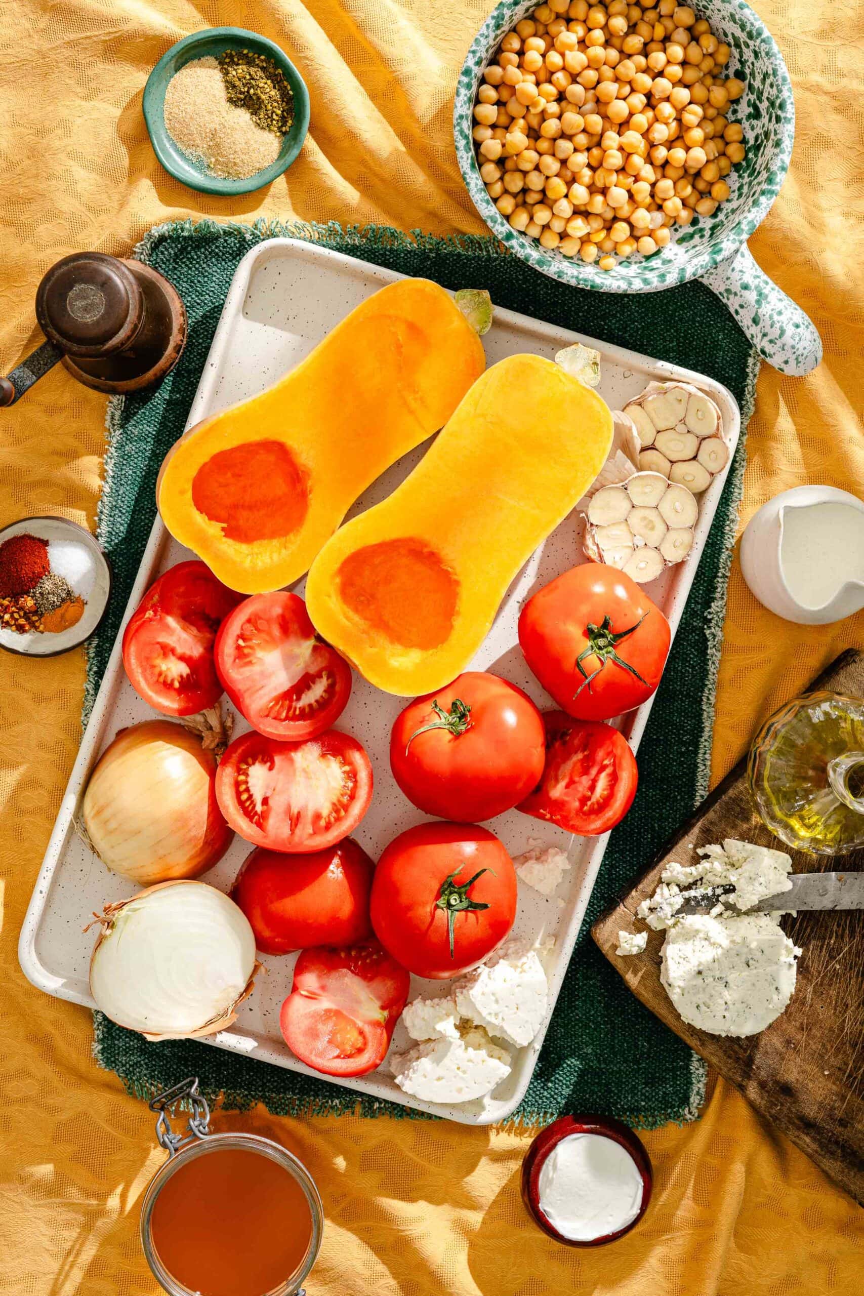 A rectangular tray holds halved butternut squash, tomatoes, and garlic cloves, surrounded by bowls of chickpeas, spices, oil, and cheese on a yellow tablecloth.