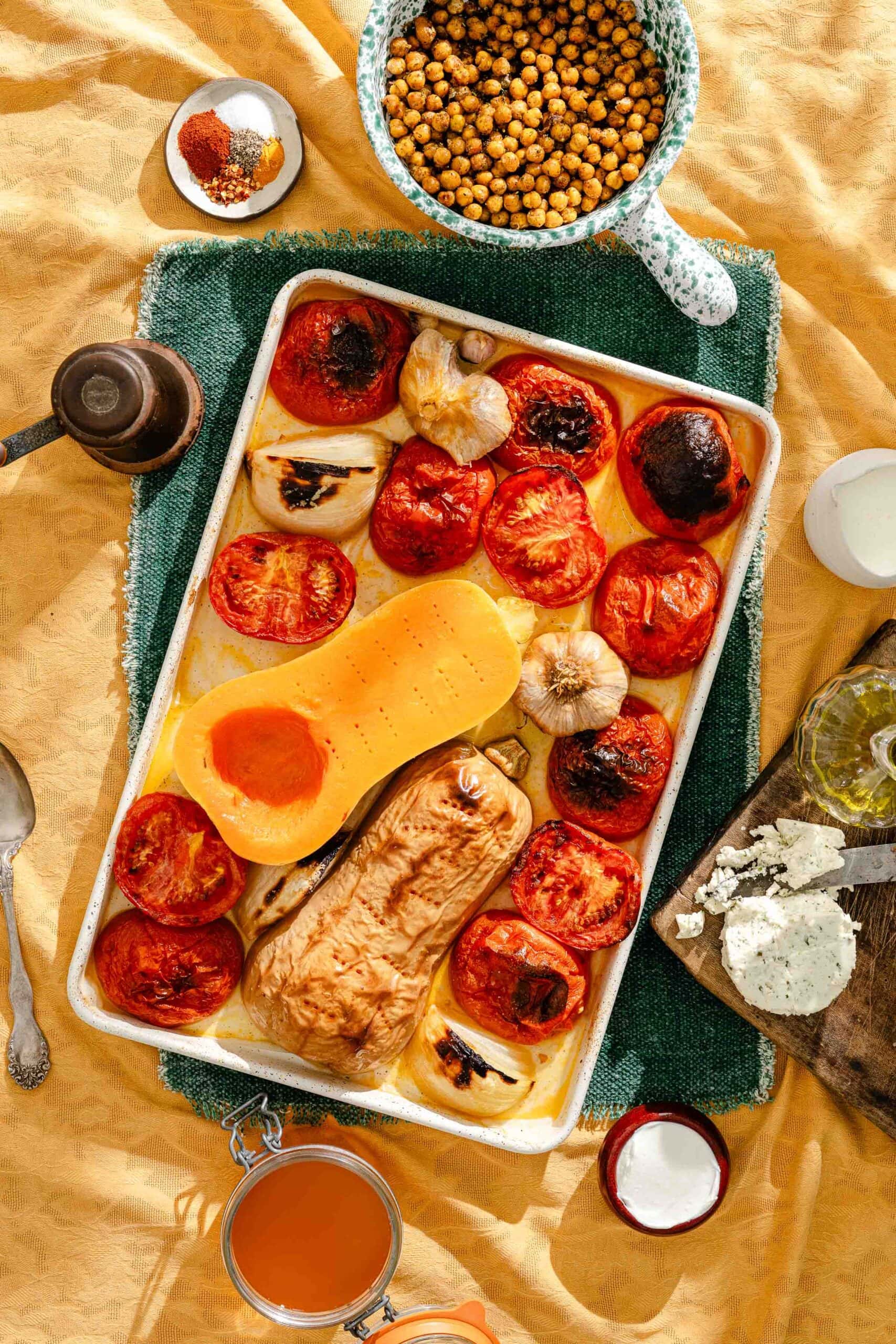 A sheet pan filled with roasted butternut squash, tomatoes, and garlic, surrounded by small bowls of chickpeas, herbs, olive oil, and crumbled cheese on a yellow tablecloth.