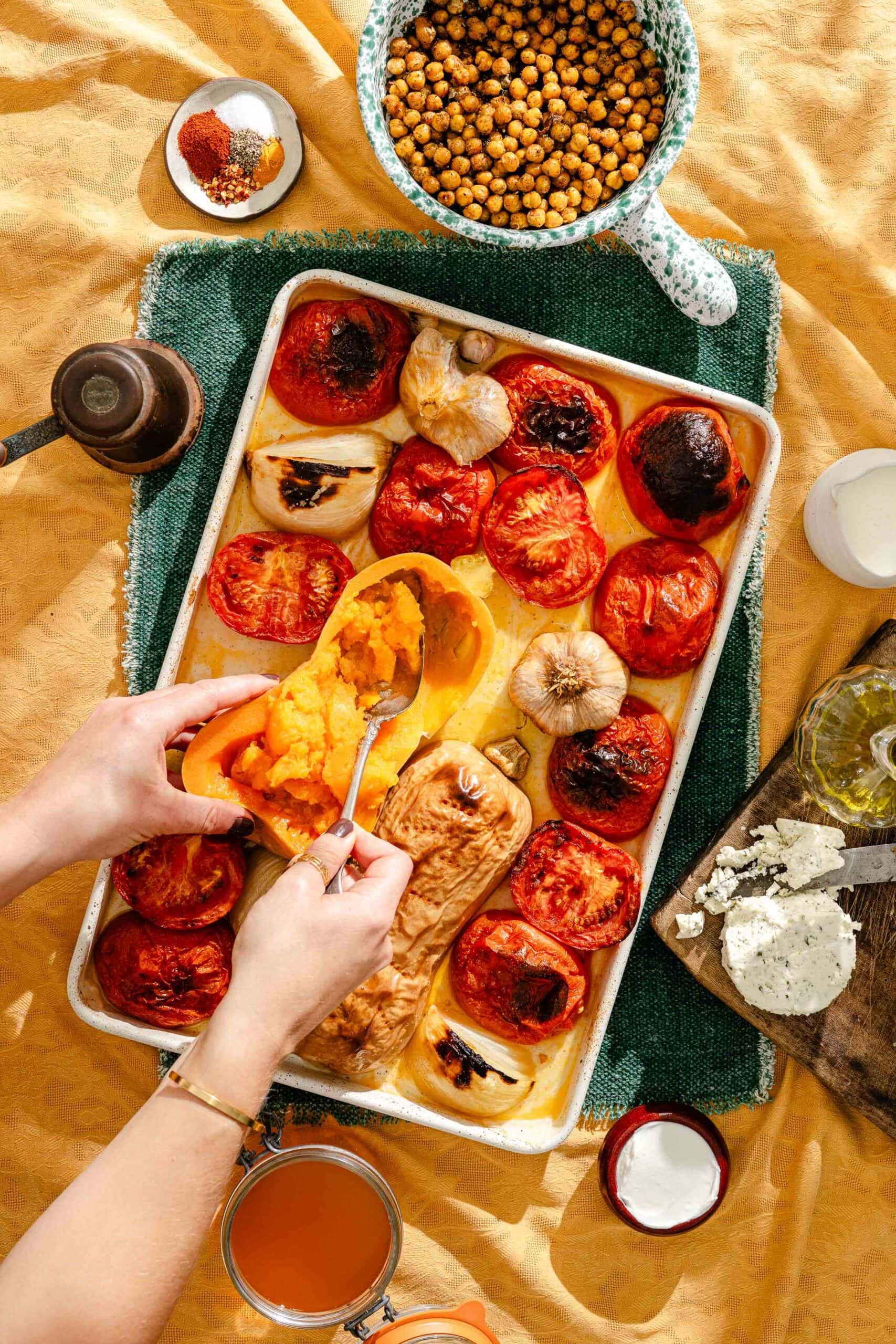 Hands scooping roasted squash from a tray of roasted tomatoes, onions, and garlic, surrounded by bowls of chickpeas, spices, and cheese.