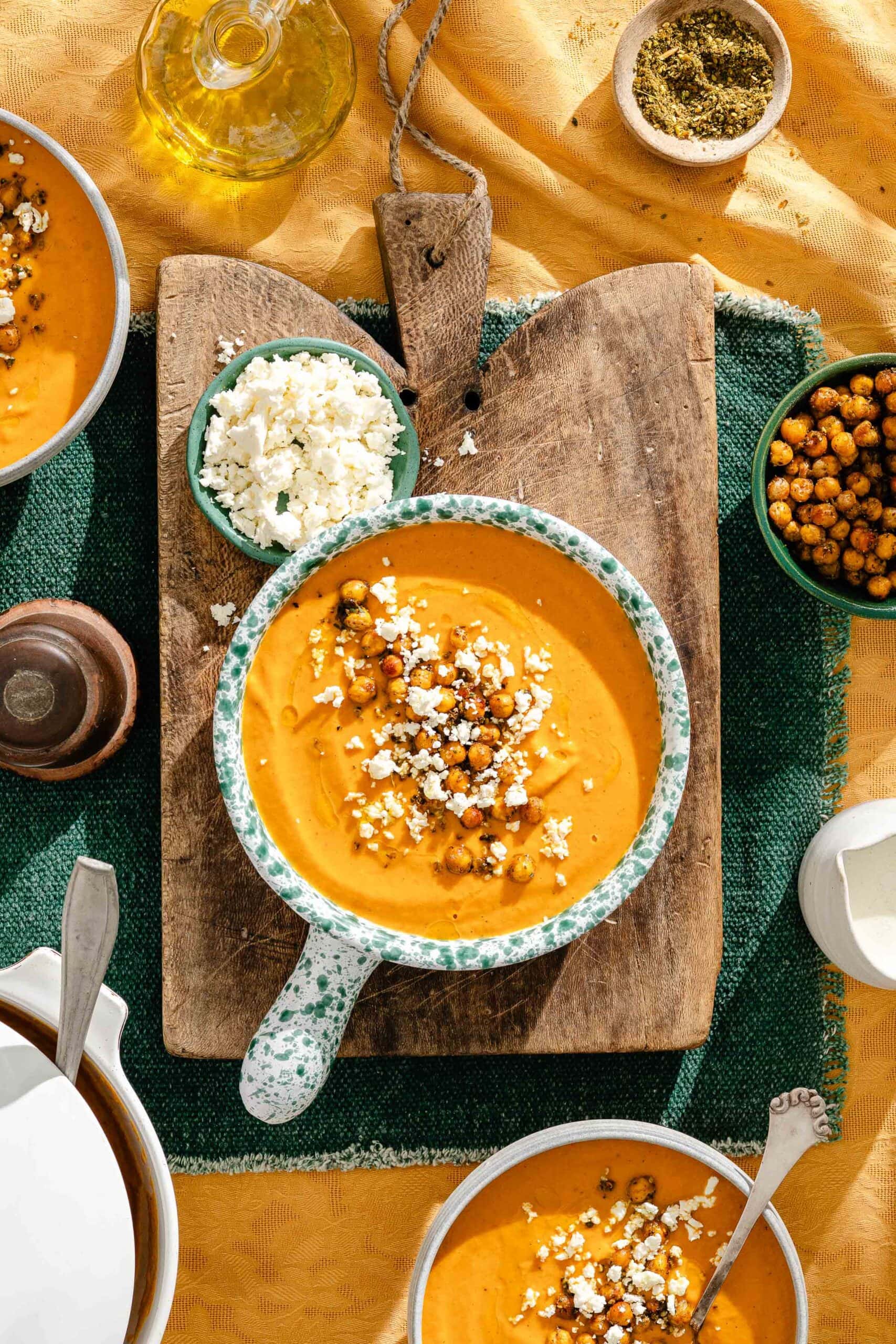 A bowl of orange soup topped with roasted chickpeas and crumbled feta, resting on a wooden board alongside bowls of additional chickpeas and feta.