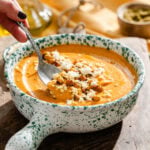 A hand uses a spoon to scoop creamy orange soup topped with roasted chickpeas and crumbled cheese from a speckled bowl.