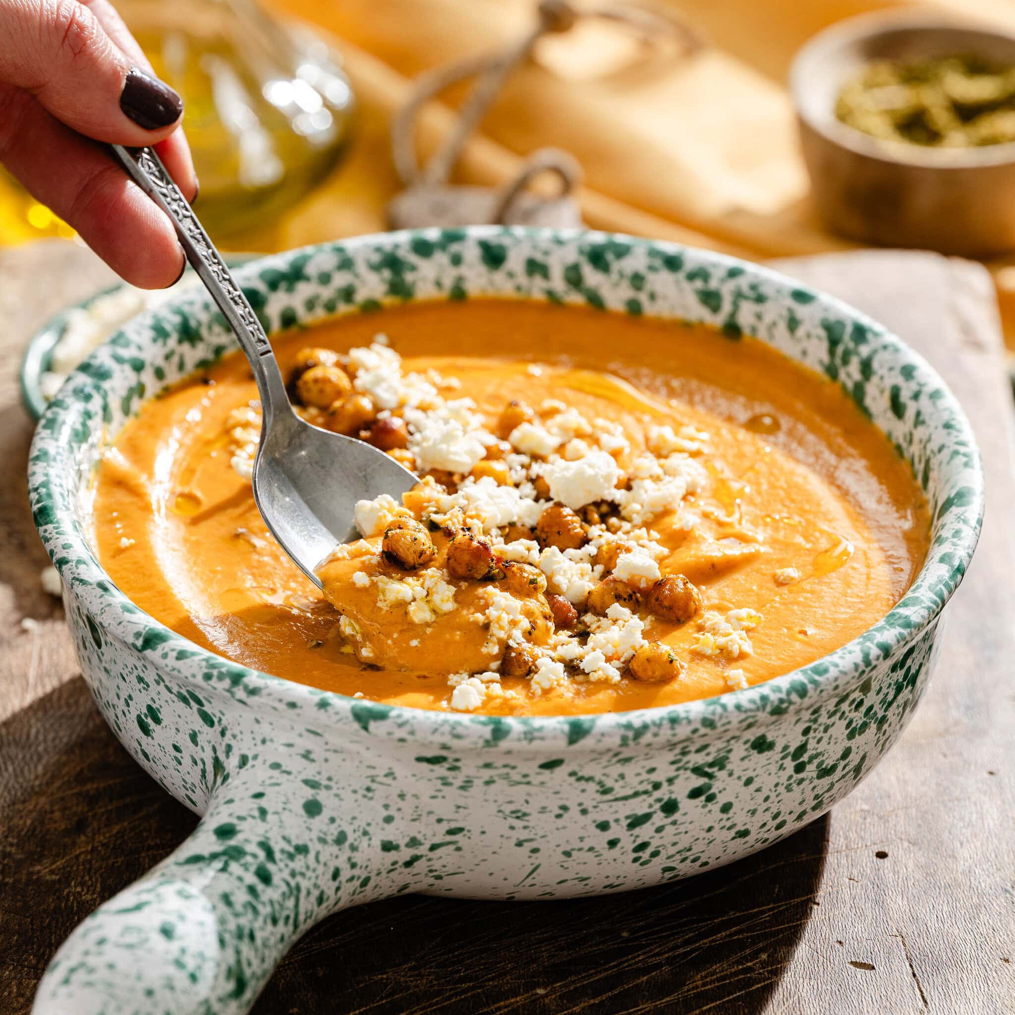 A hand uses a spoon to scoop creamy orange soup topped with roasted chickpeas and crumbled cheese from a speckled bowl.