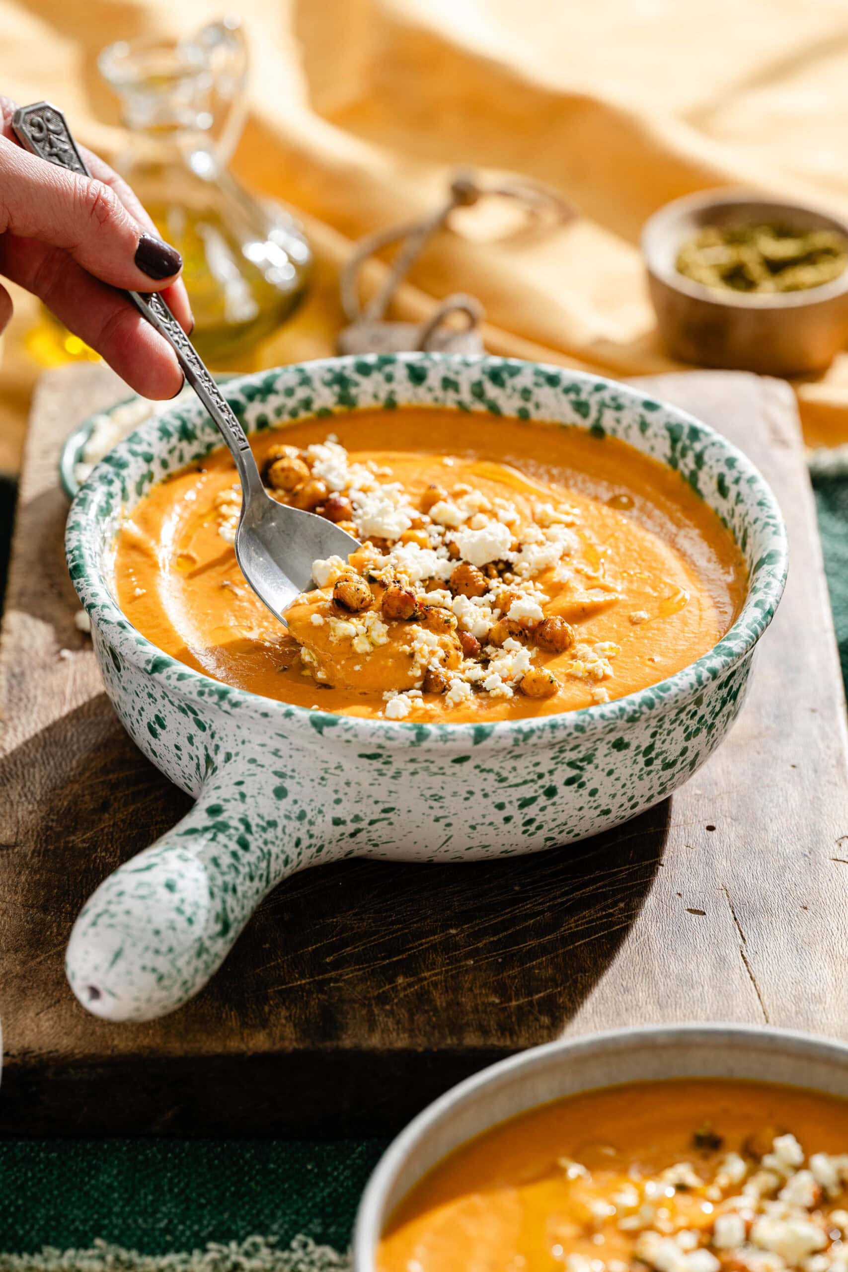A hand holding a spoon dips into a bowl of orange soup topped with roasted chickpeas and crumbled cheese.