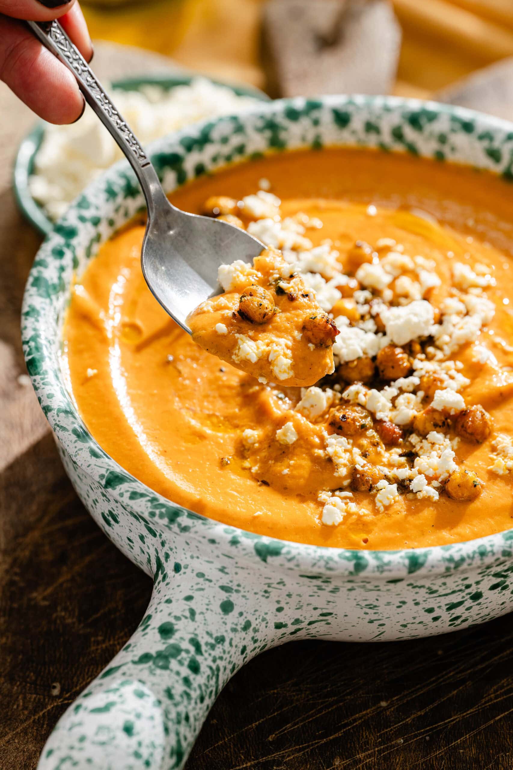 A spoon lifts a portion of creamy orange soup topped with roasted chickpeas and crumbled feta from a speckled bowl.