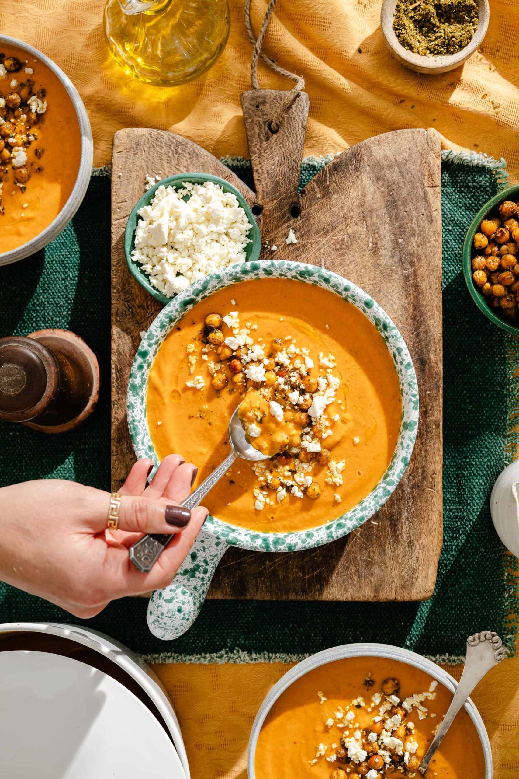 A hand holds a spoon in a bowl of creamy orange soup topped with roasted chickpeas and crumbled feta, set on a wooden board with side dishes of the same toppings.