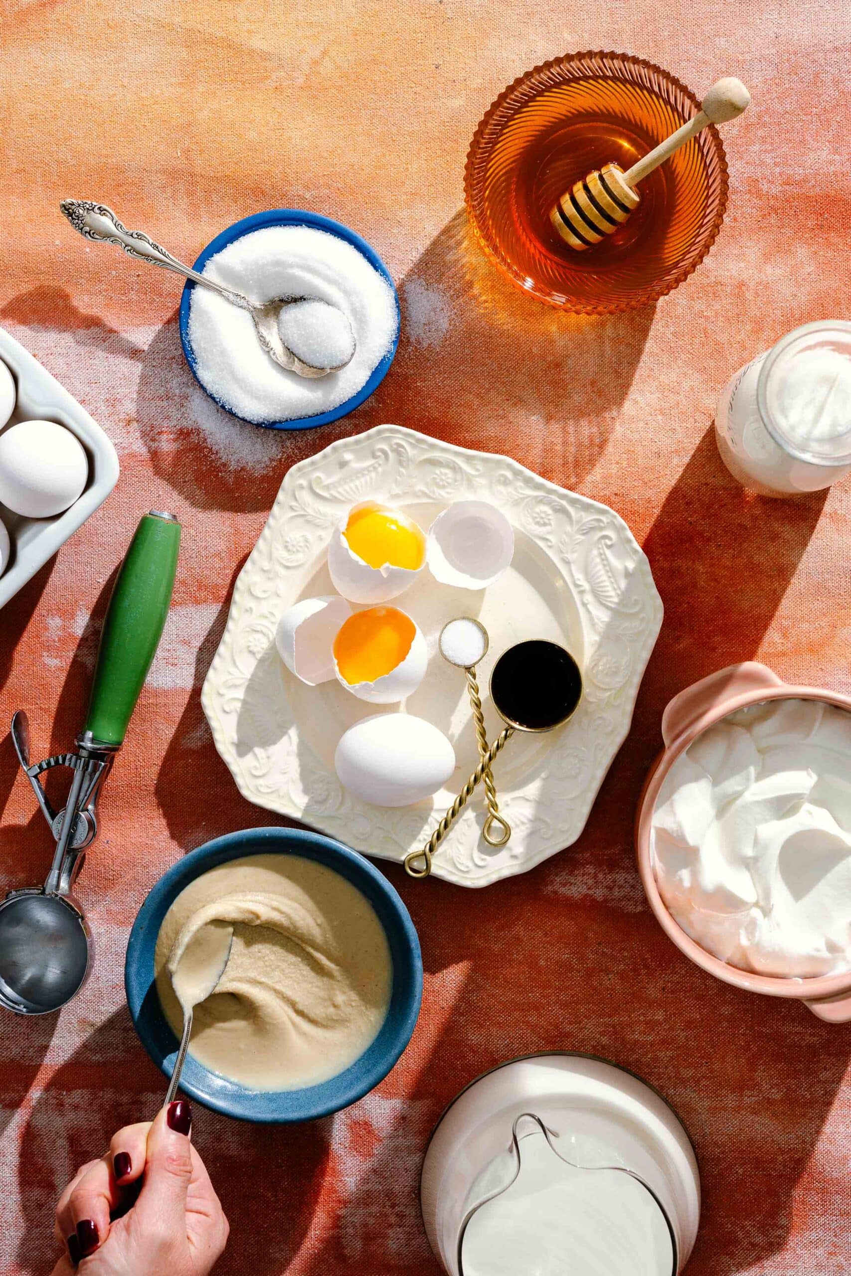 Various baking ingredients—eggs, sugar, honey, vanilla, and cream—are arranged on a colorful tabletop with bowls, a spoon, and a hand mixing batter in a blue bowl.