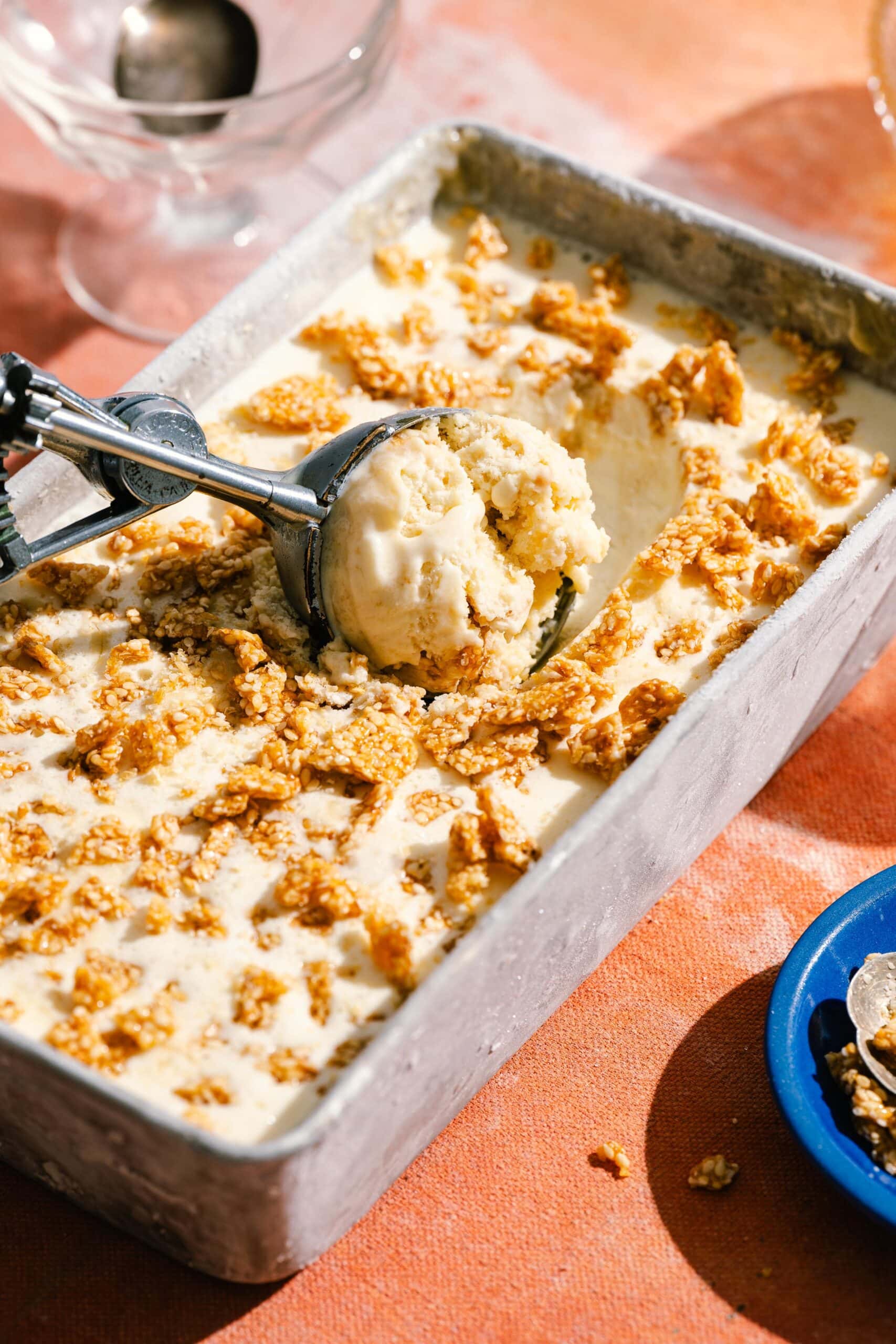 A metal pan filled with creamy ice cream topped with golden crunchy flakes. An ice cream scooper holds a scoop above the dessert. The pan is on an orange surface, with a blue dish and utensil nearby.