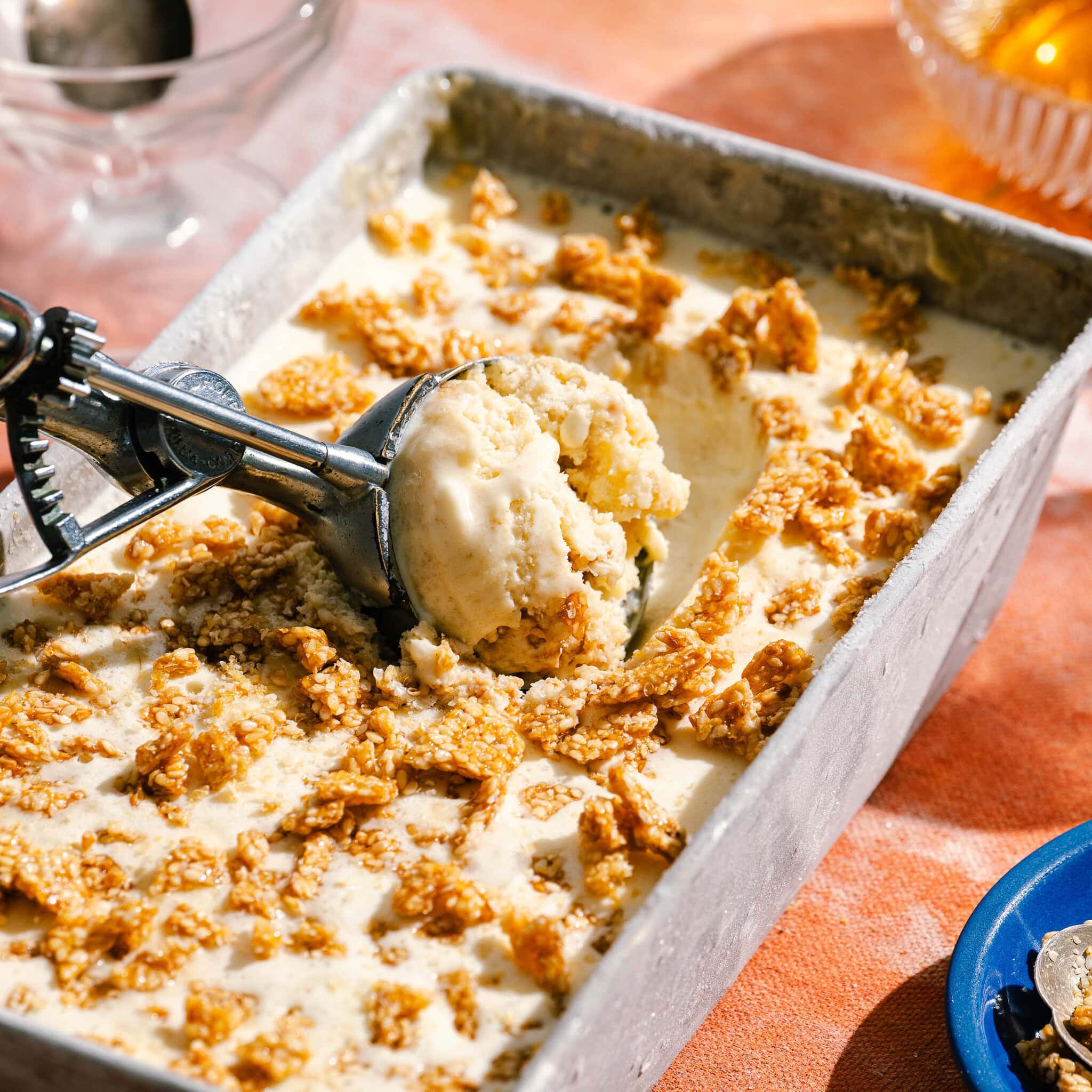 A metal pan of creamy ice cream topped with crunchy cereal, with an ice cream scoop lifting a serving. The pan sits on a peach-colored surface, with a blue plate and dishes in the background.