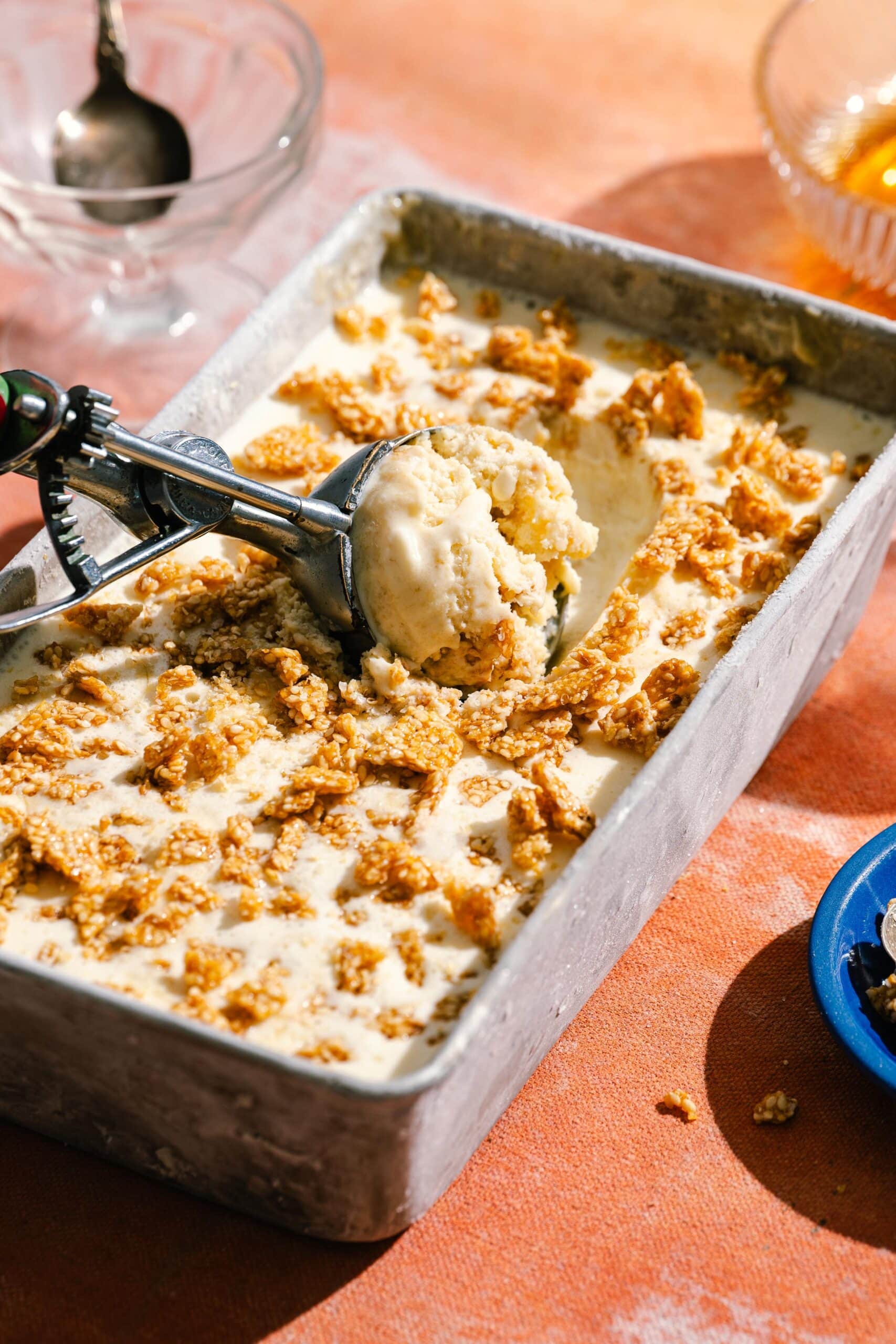 A metal tray of ice cream topped with crunchy cereal flakes is being scooped with an ice cream scooper. The scene is set on an orange surface with bowls and honey nearby.