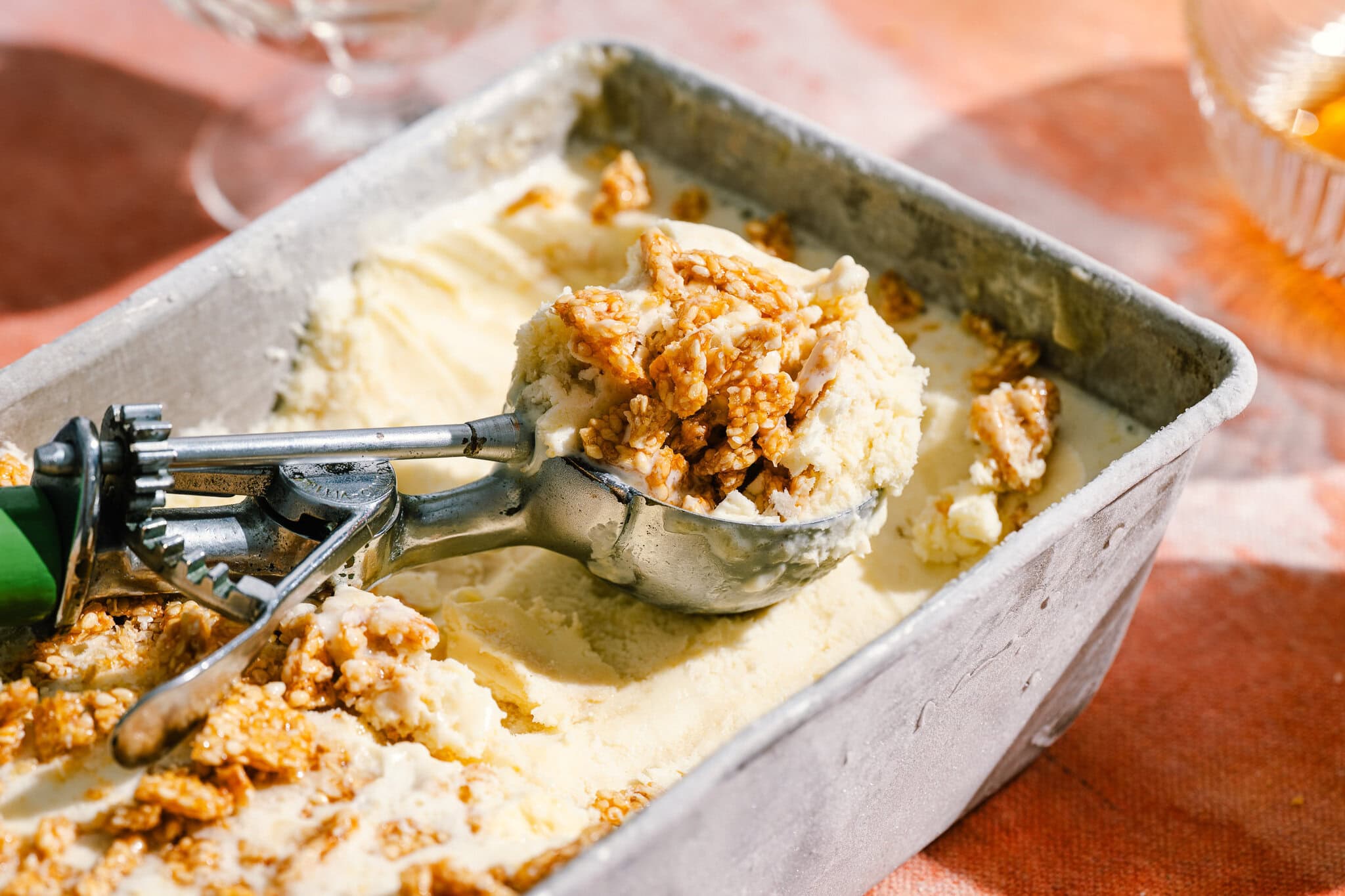 An ice cream scoop lifting a serving of vanilla ice cream topped with crunchy granola from a metal container, on a peach-colored tablecloth.