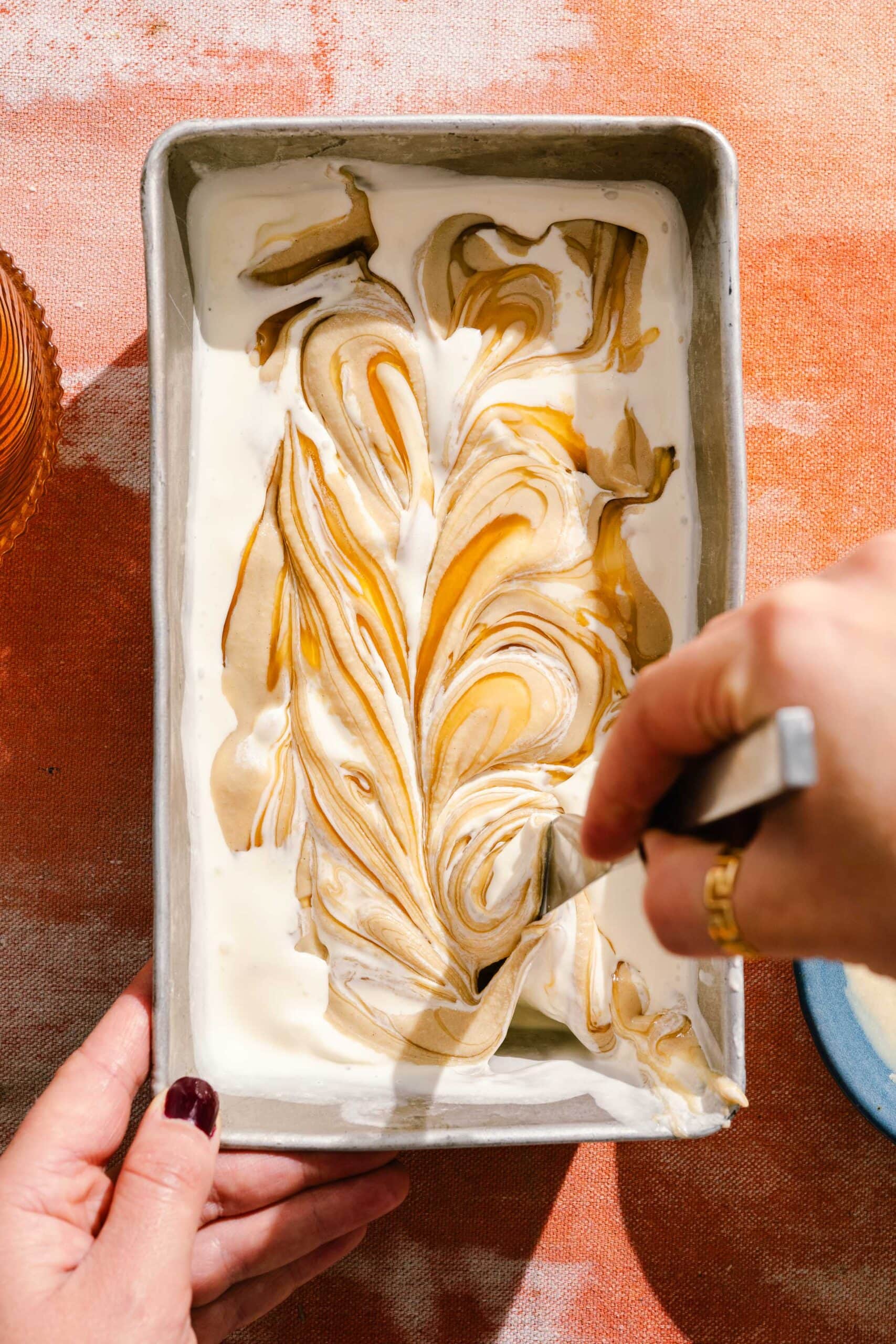 A person swirls caramel into a pan of creamy white ice cream with a spoon, creating a marbled pattern.