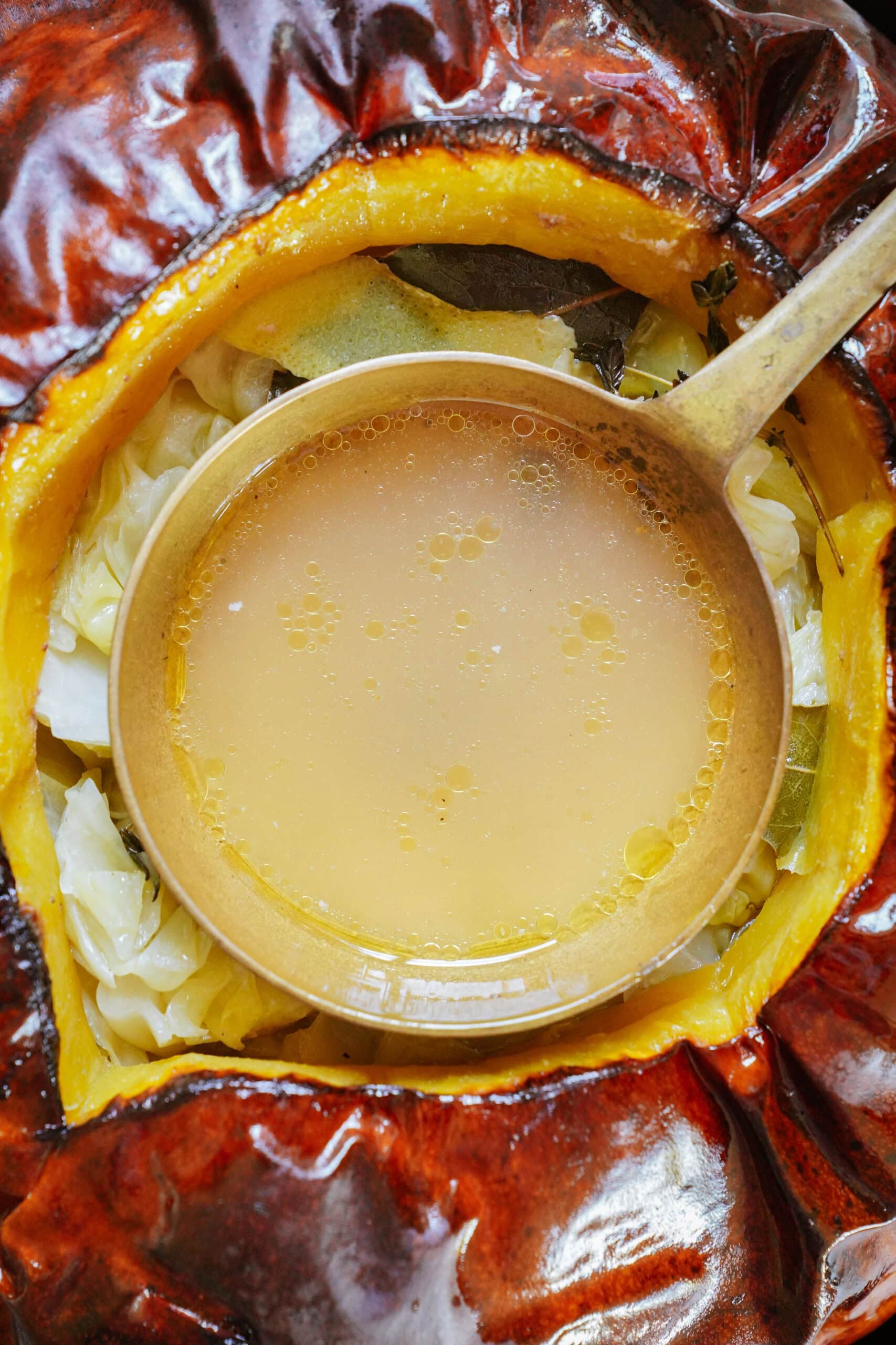 A ladle filled with broth rests inside a cooked pumpkin, surrounded by pieces of cabbage and herbs.