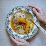 A person holds a patterned bowl of soup containing chicken, vegetables, and herbs, with a spoon resting on the edge. The bowl sits on a light-colored surface.