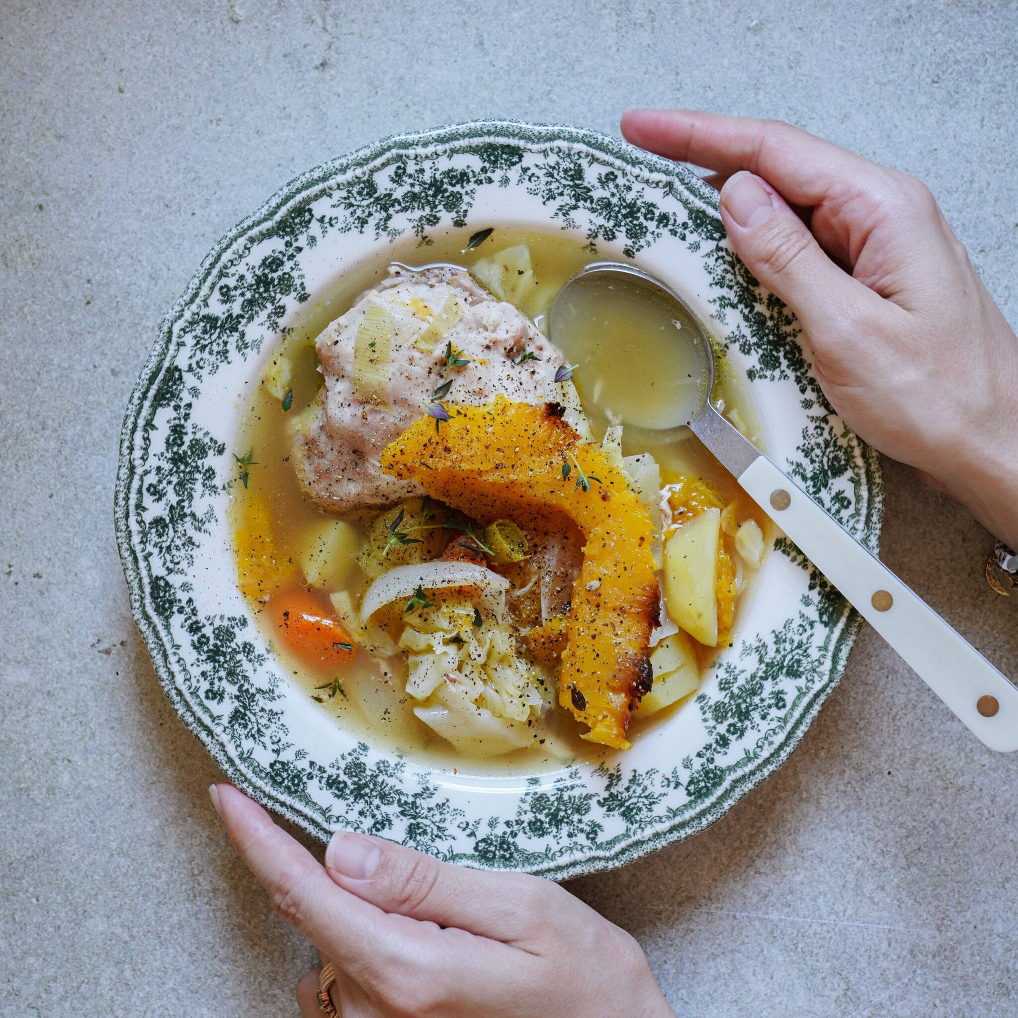 A person holds a patterned bowl of soup containing chicken, vegetables, and herbs, with a spoon resting on the edge. The bowl sits on a light-colored surface.
