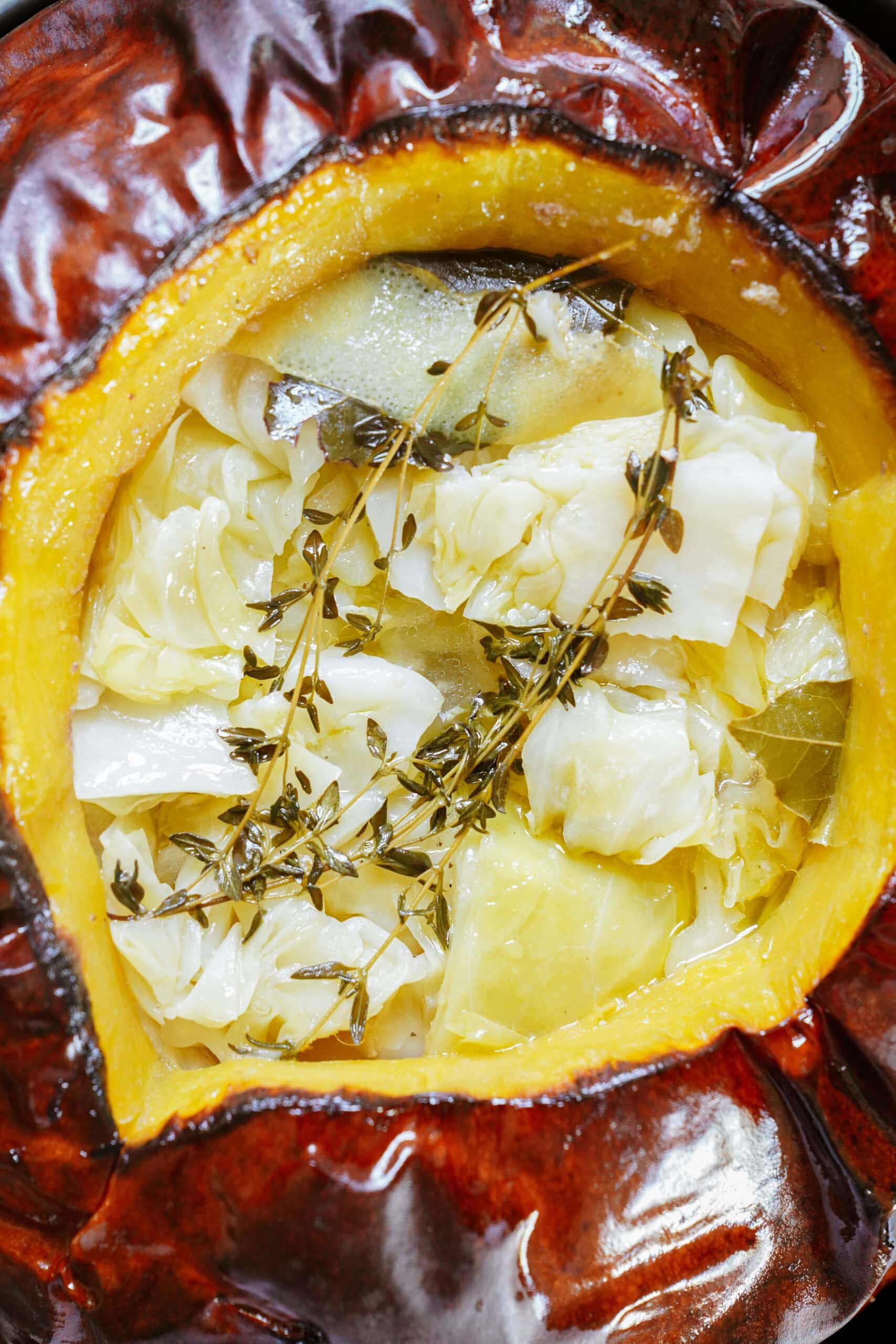 A roasted pumpkin filled with cooked cabbage, herbs, and bay leaves, viewed from above. The pumpkins orange interior and glossy brown skin contrast with the light-colored cabbage.