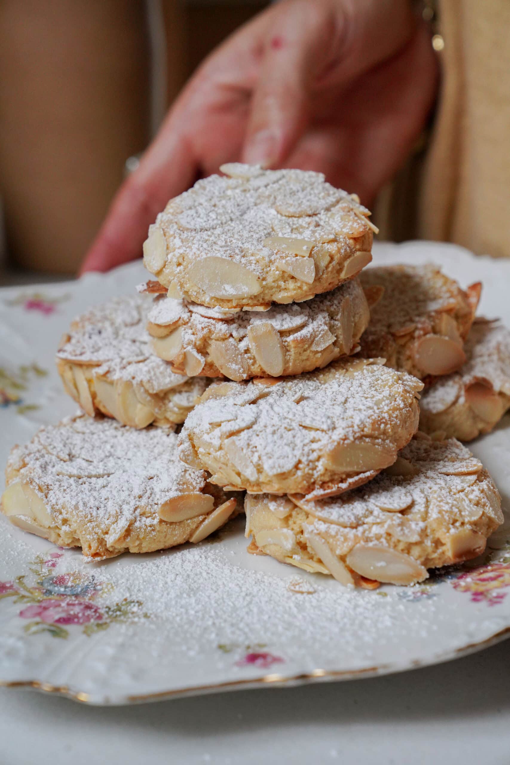 A stack of almond cookies dusted with powdered sugar on a floral china plate.