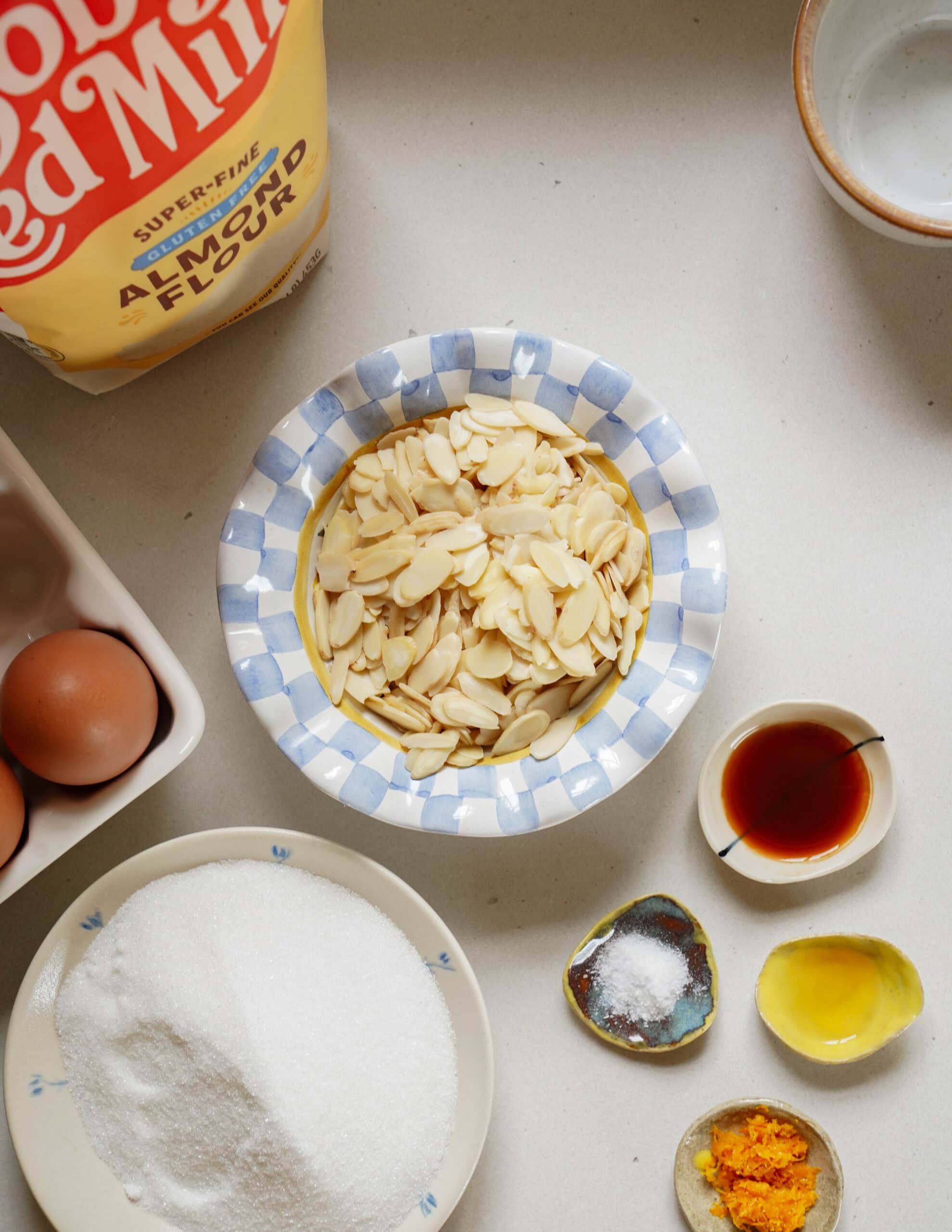 Ingredients for a recipe arranged on a counter, including a bag of almond flour, a bowl of sliced almonds, sugar, eggs, vanilla extract, salt, and orange zest.
