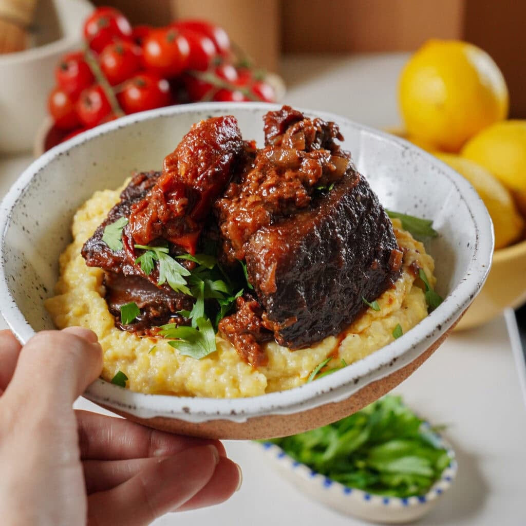 A hand holds a ceramic bowl filled with polenta topped with braised short rib and fresh herbs.