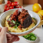 A hand holds a ceramic bowl filled with polenta topped with braised short rib and fresh herbs.