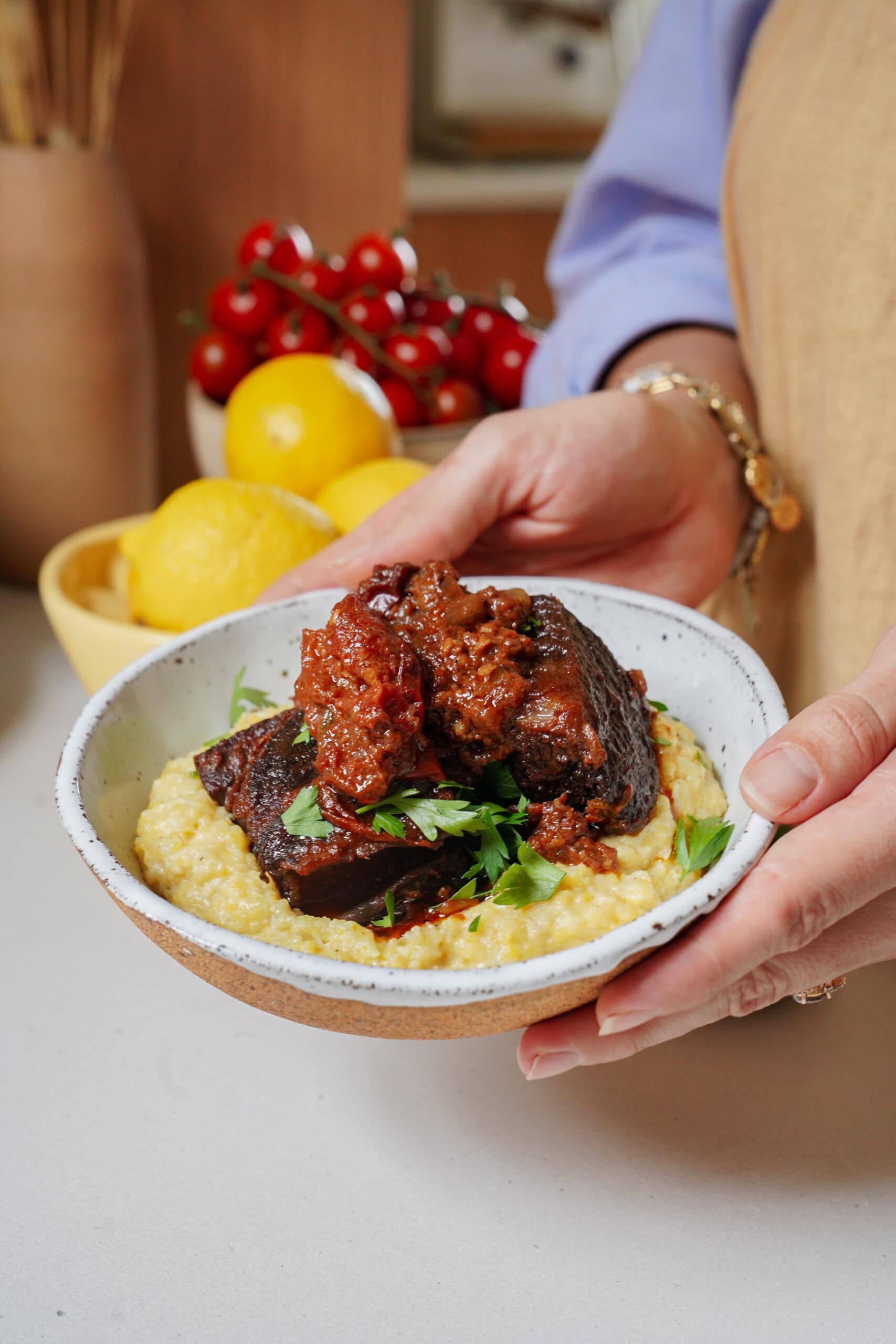 A person holding a bowl of braised short ribs served over polenta and garnished with parsley.