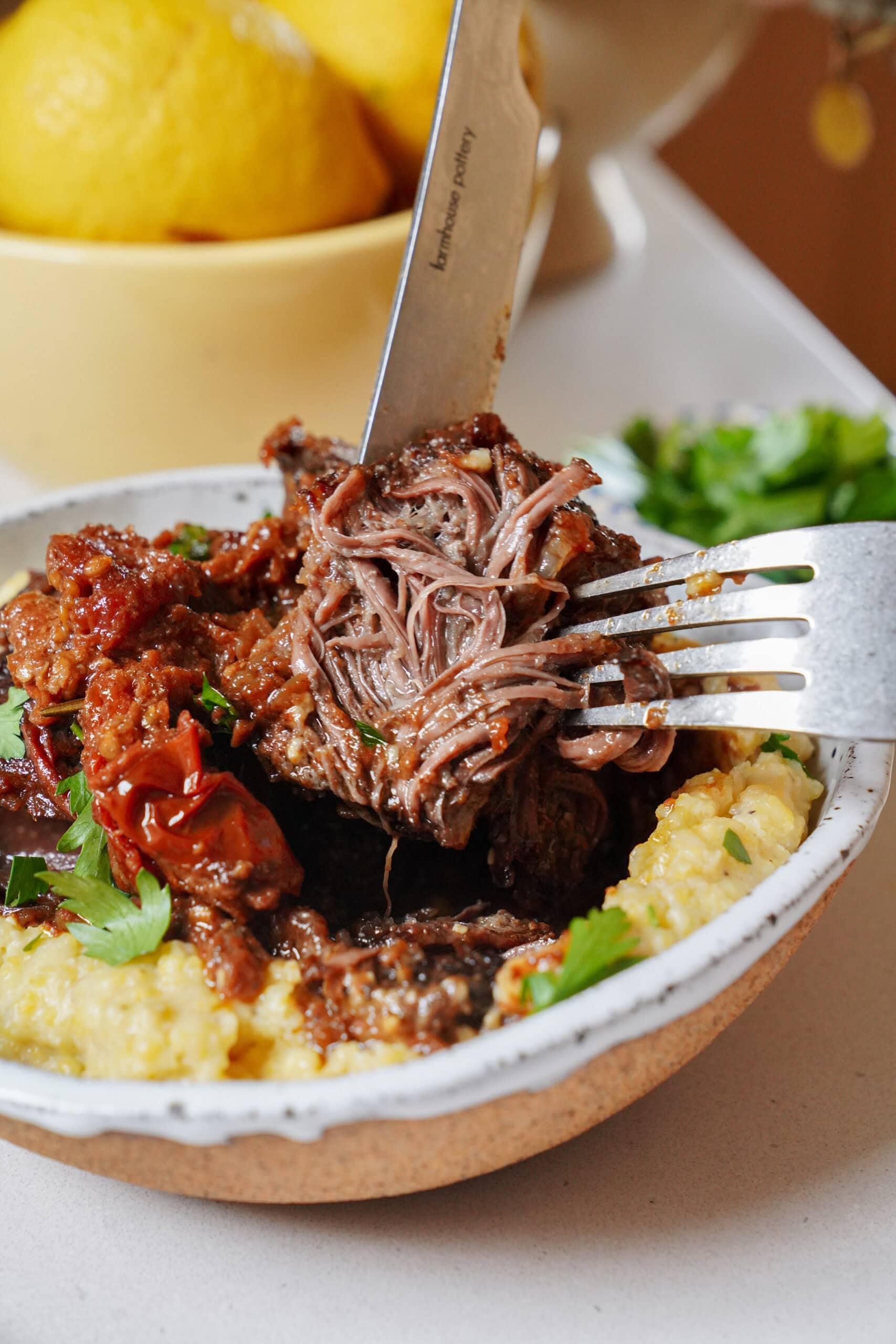A knife and fork lift a piece of shredded, braised beef with tomatoes from a bowl of creamy polenta.