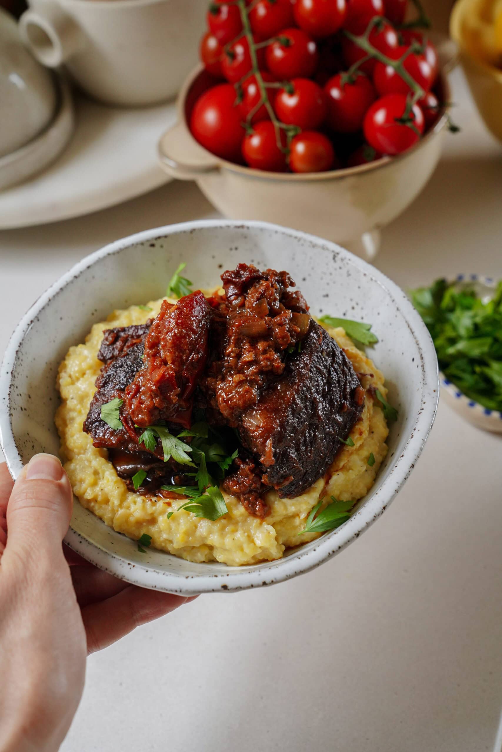A hand holding a speckled bowl of polenta topped with braised short rib and herbs.