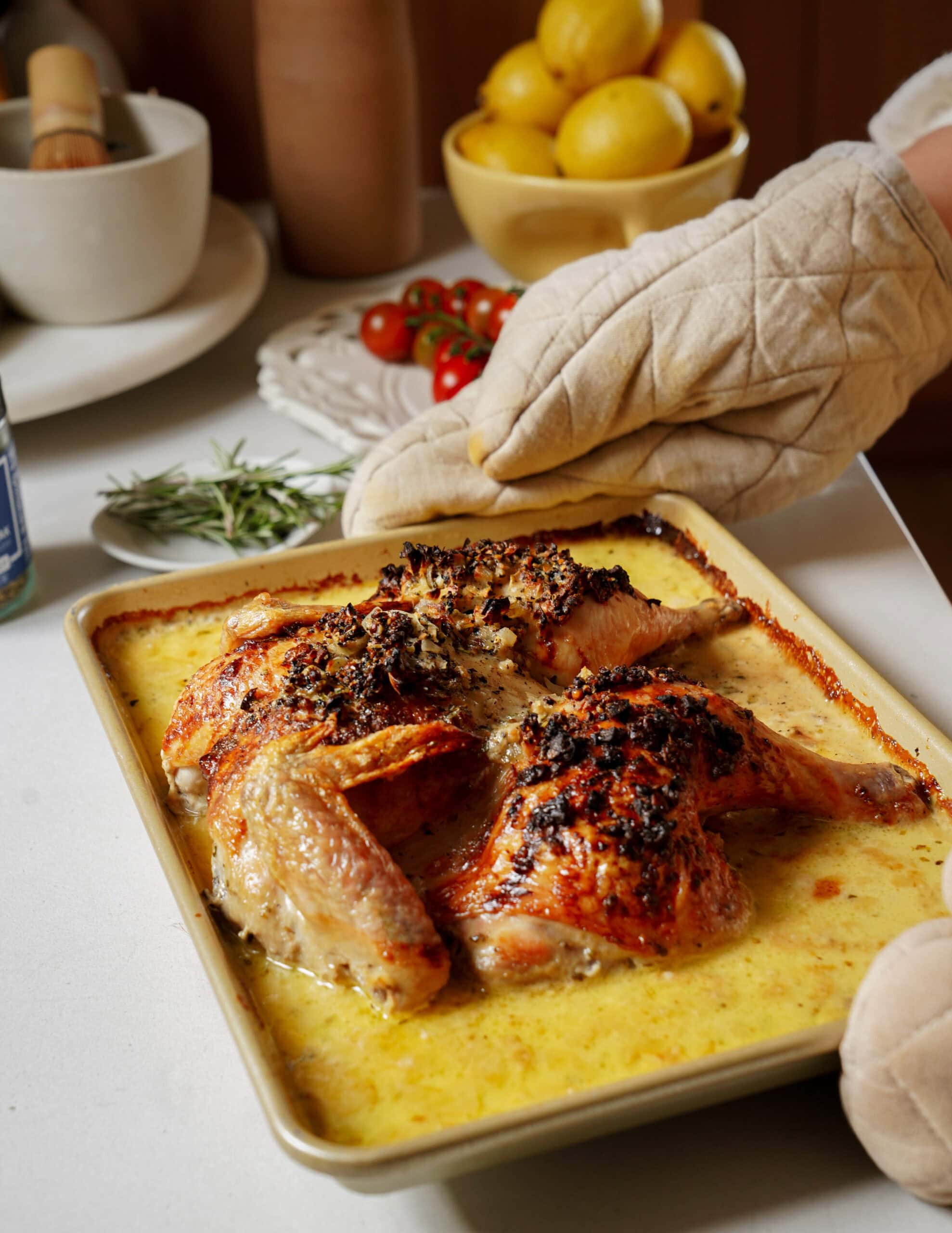 A person wearing an oven mitt holds a baking dish containing a roasted chicken in a creamy sauce.