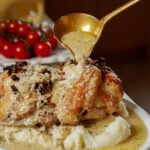 A gold ladle pours a creamy sauce over a roasted chicken served with mashed potatoes, with a cluster of cherry tomatoes in the background.