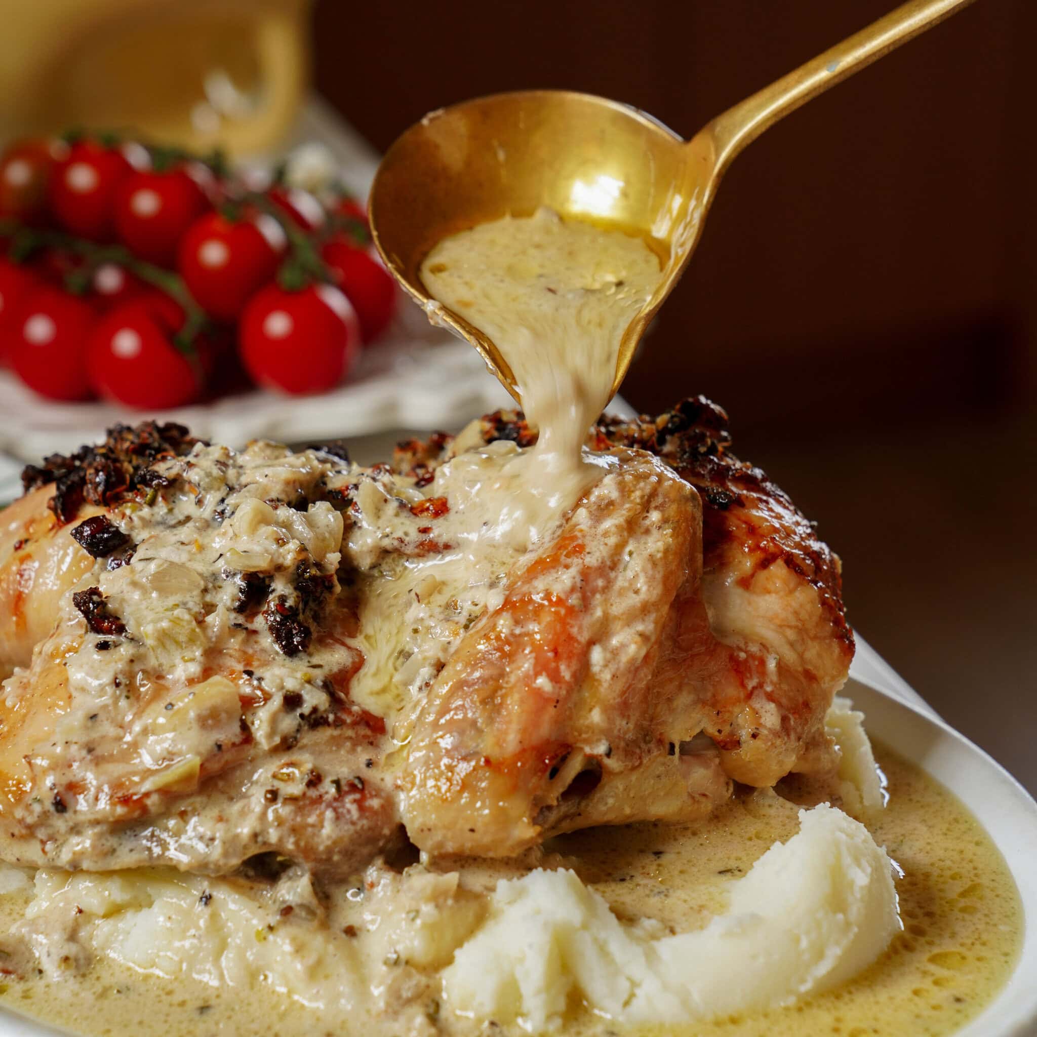 A gold ladle pours a creamy sauce over a roasted chicken served with mashed potatoes, with a cluster of cherry tomatoes in the background.