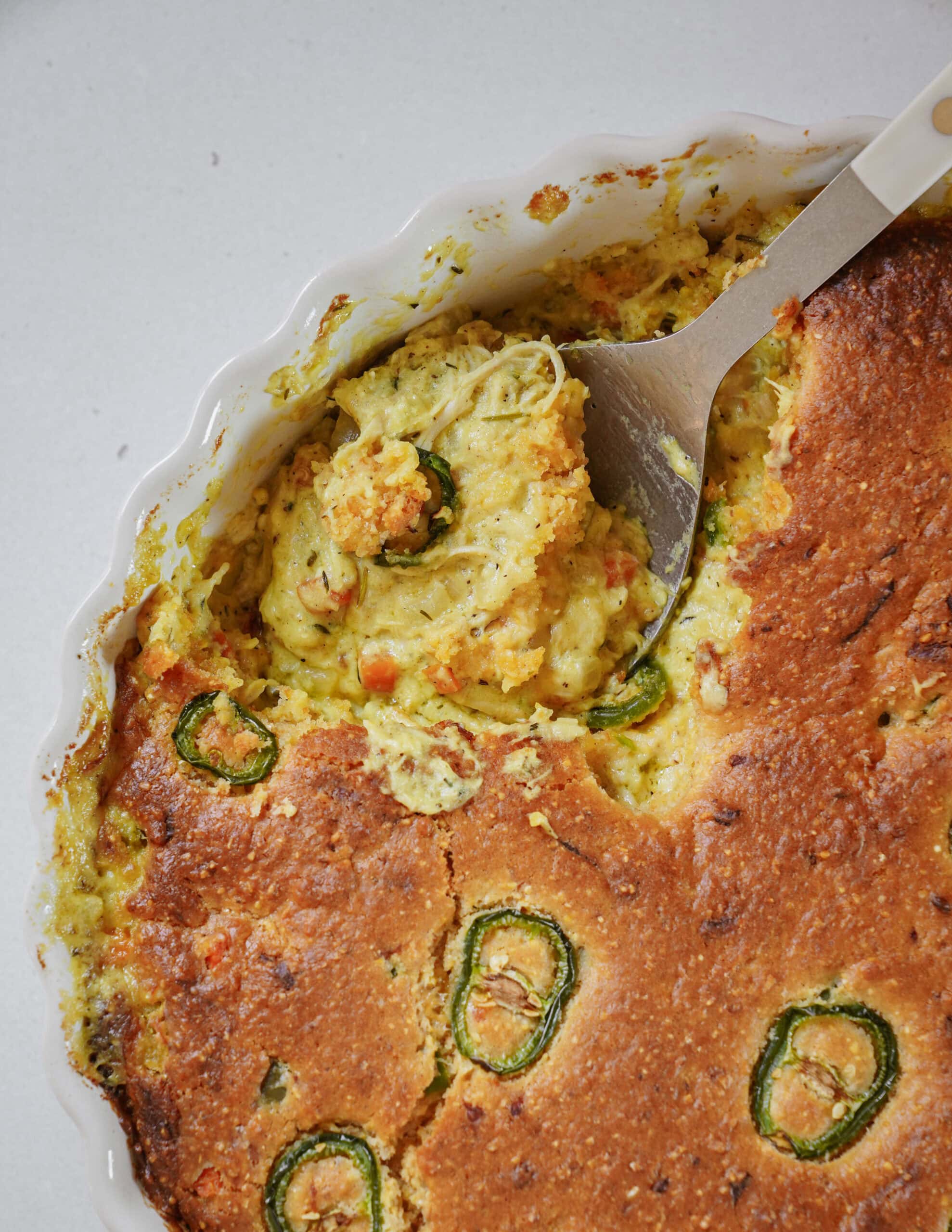 A serving spoon lifting a scoop of savory corn casserole topped with sliced jalapeños from a scalloped baking dish.
