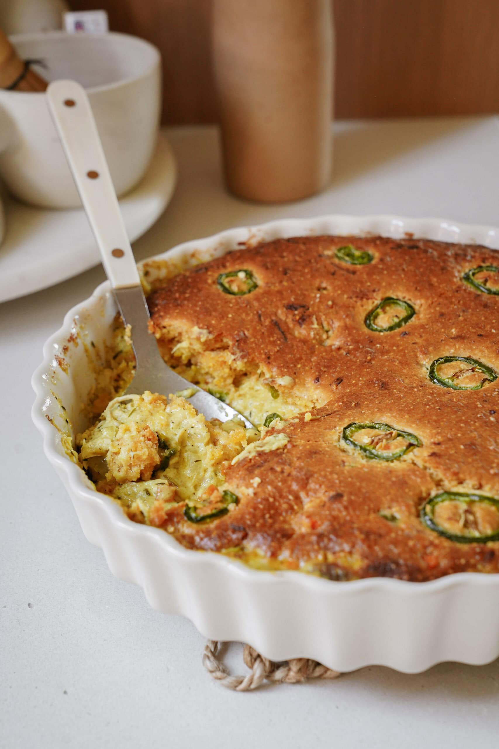 A scalloped baking dish containing jalapeño cornbread, with a serving spoon lifting a portion.