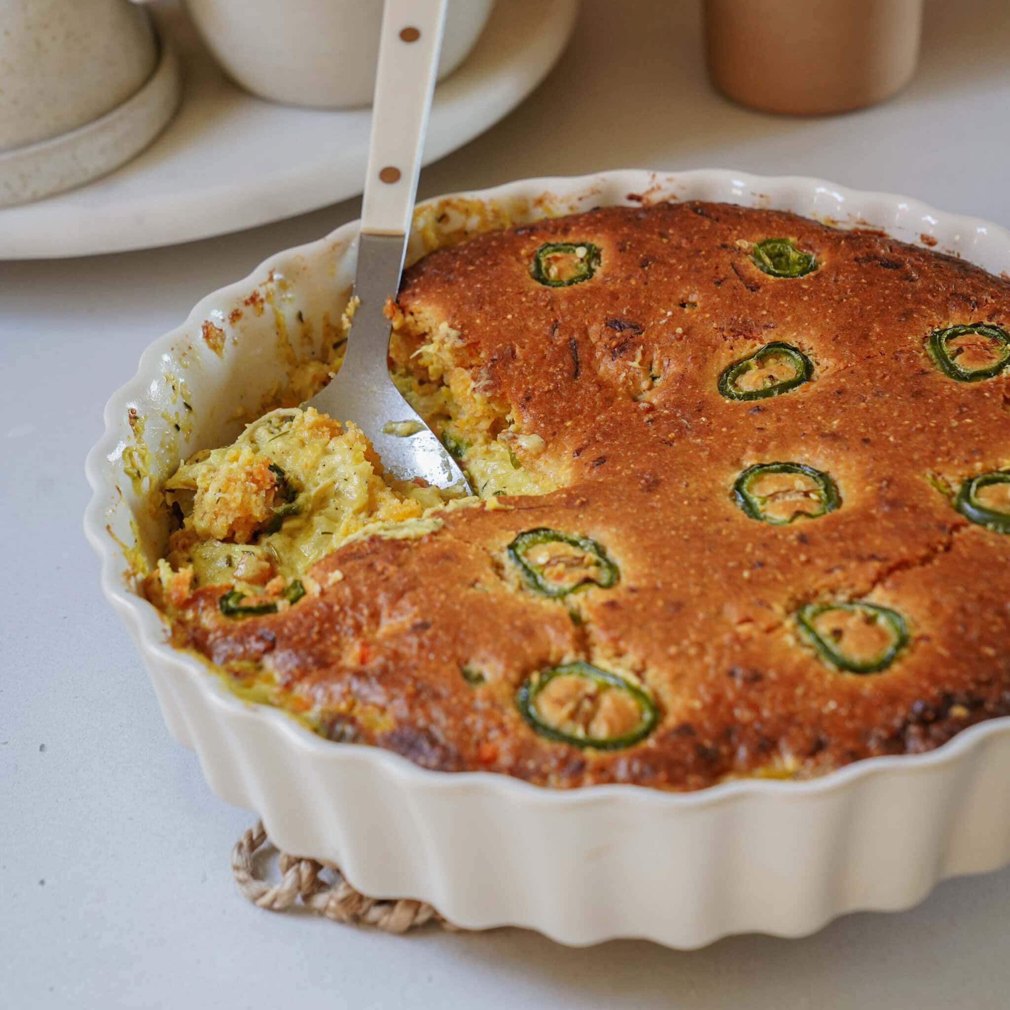 A baked cornbread casserole topped with jalapeño slices in a white fluted dish, with a serving spoon resting inside.