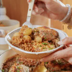 A person holds a bowl containing chicken thighs and pearl onions in a savory sauce, lifting a portion with a fork.