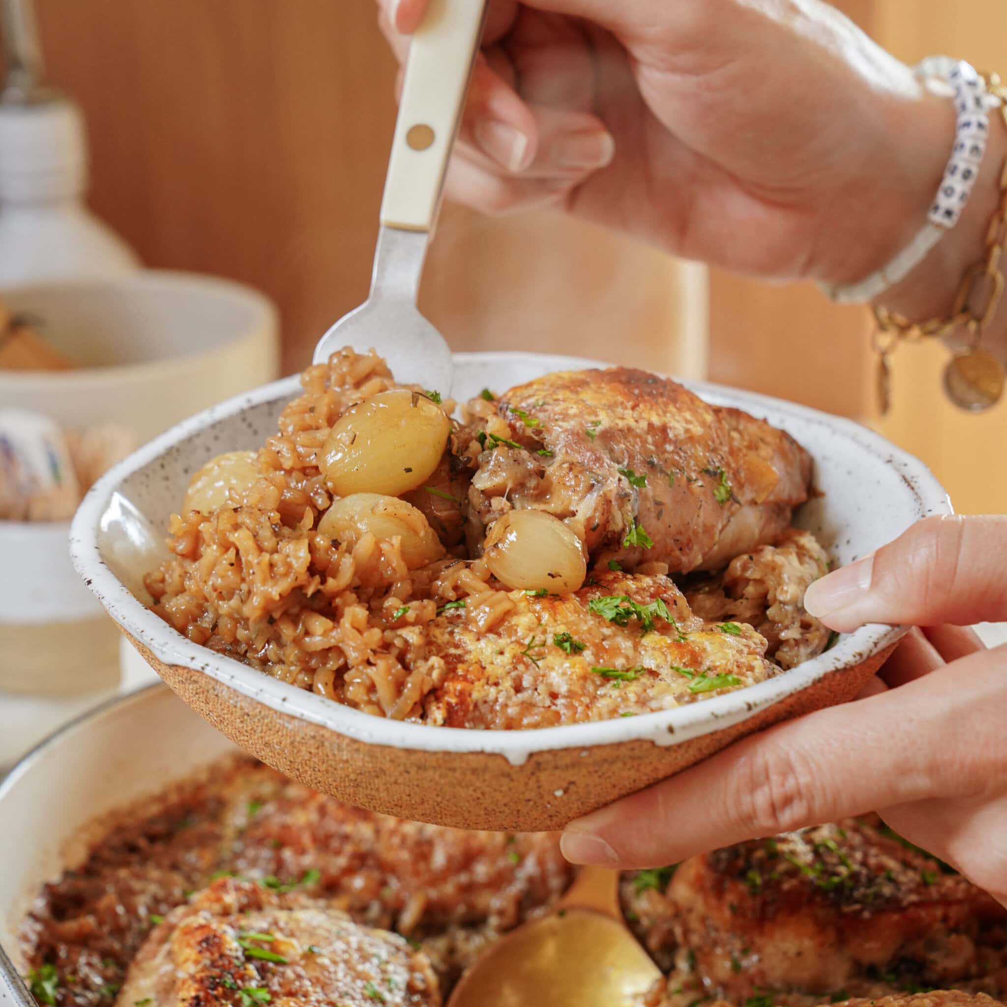 A person holds a bowl containing chicken thighs and pearl onions in a savory sauce, lifting a portion with a fork.