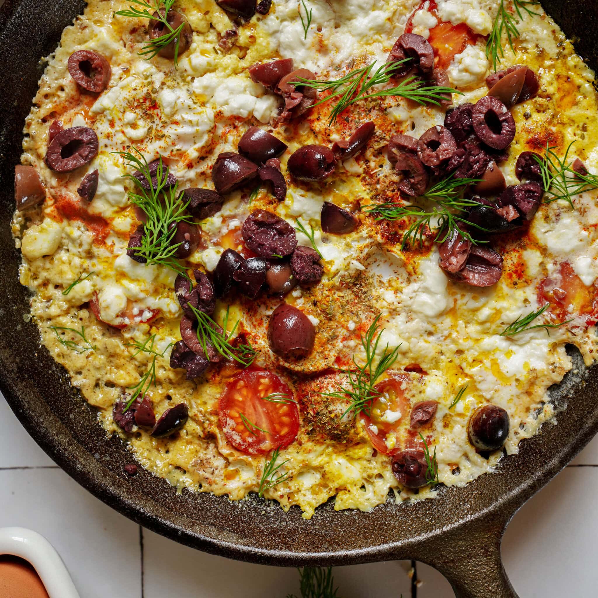 Cast iron skillet containing a cooked egg scramble with sliced cherry tomatoes, crumbled white cheese, pitted black olives, and fresh dill.