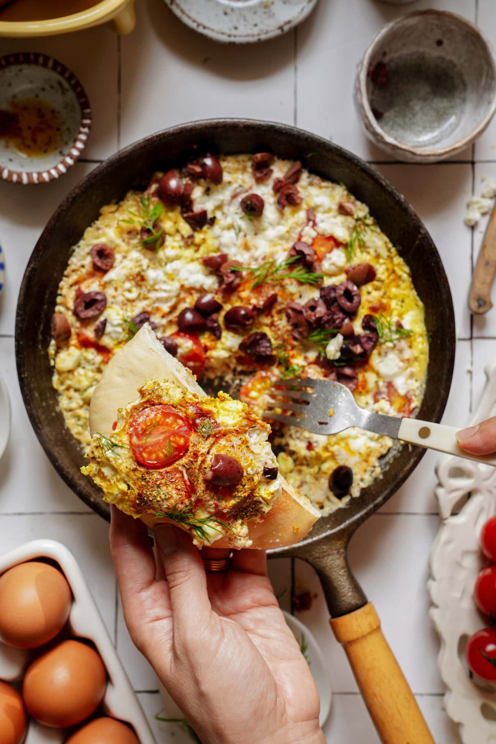 A hand holds a piece of pita bread topped with a scoop of egg scramble containing tomatoes, feta cheese, and olives, with the remainder of the dish in a skillet below.