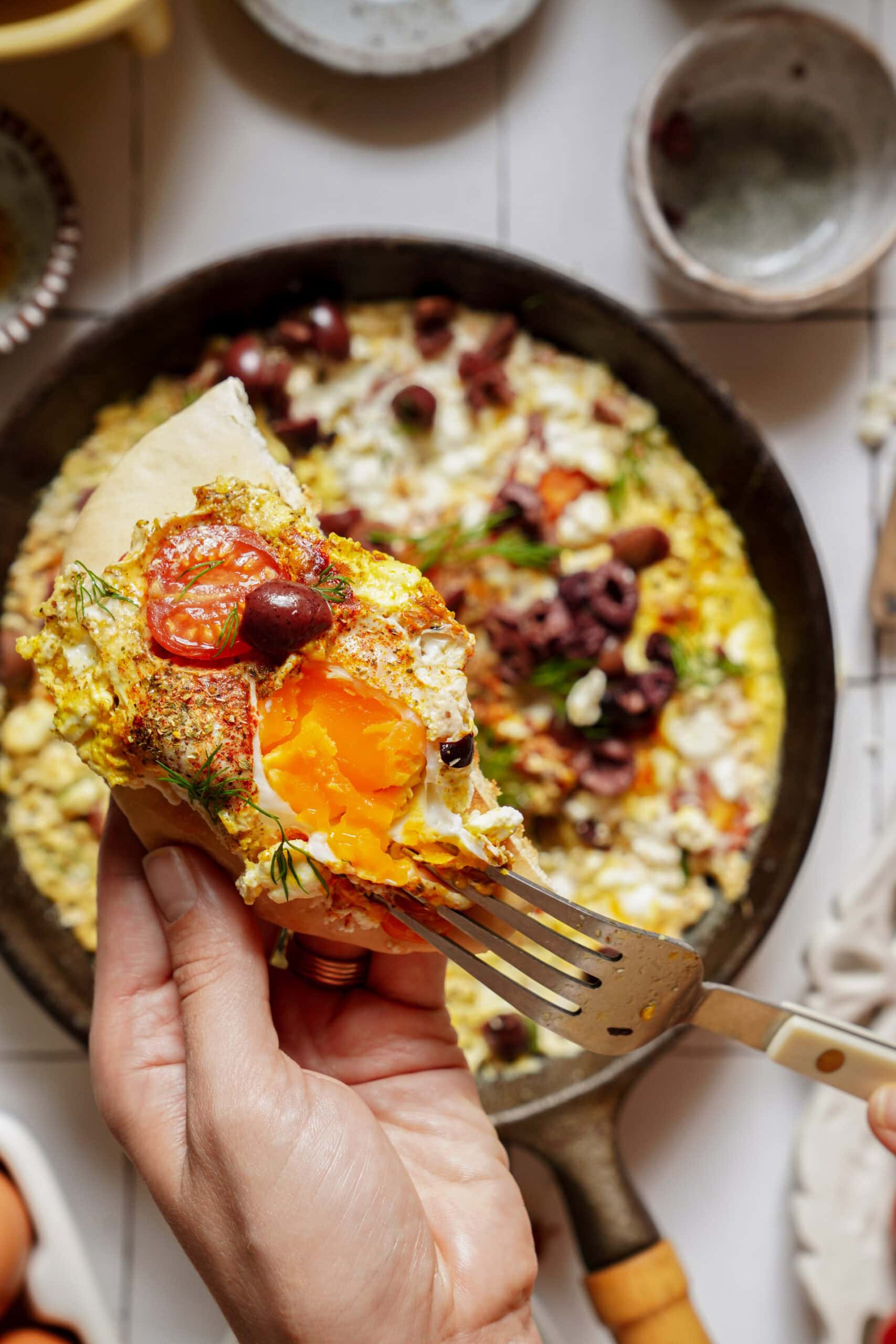 A hand holding a piece of pita topped with a fried egg, tomato, and olives, with a fork breaking into the runny yolk above a skillet of shakshuka.