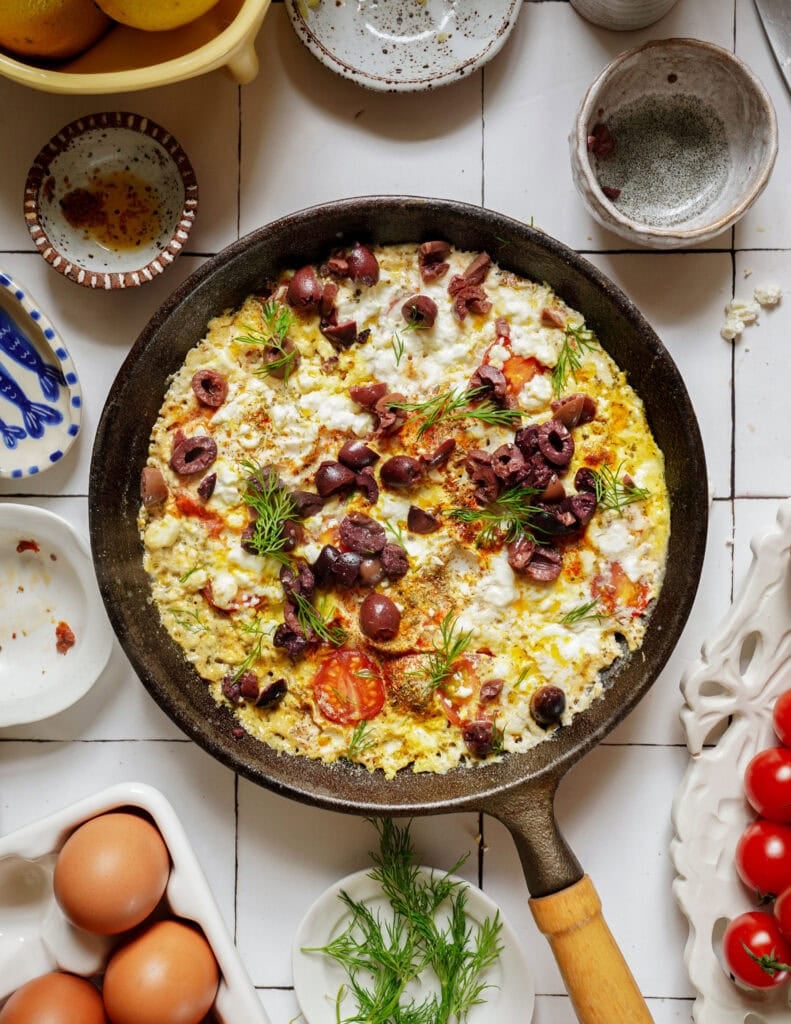 A cast iron skillet filled with a cooked egg scramble topped with feta, sliced kalamata olives, cherry tomatoes, and fresh dill.