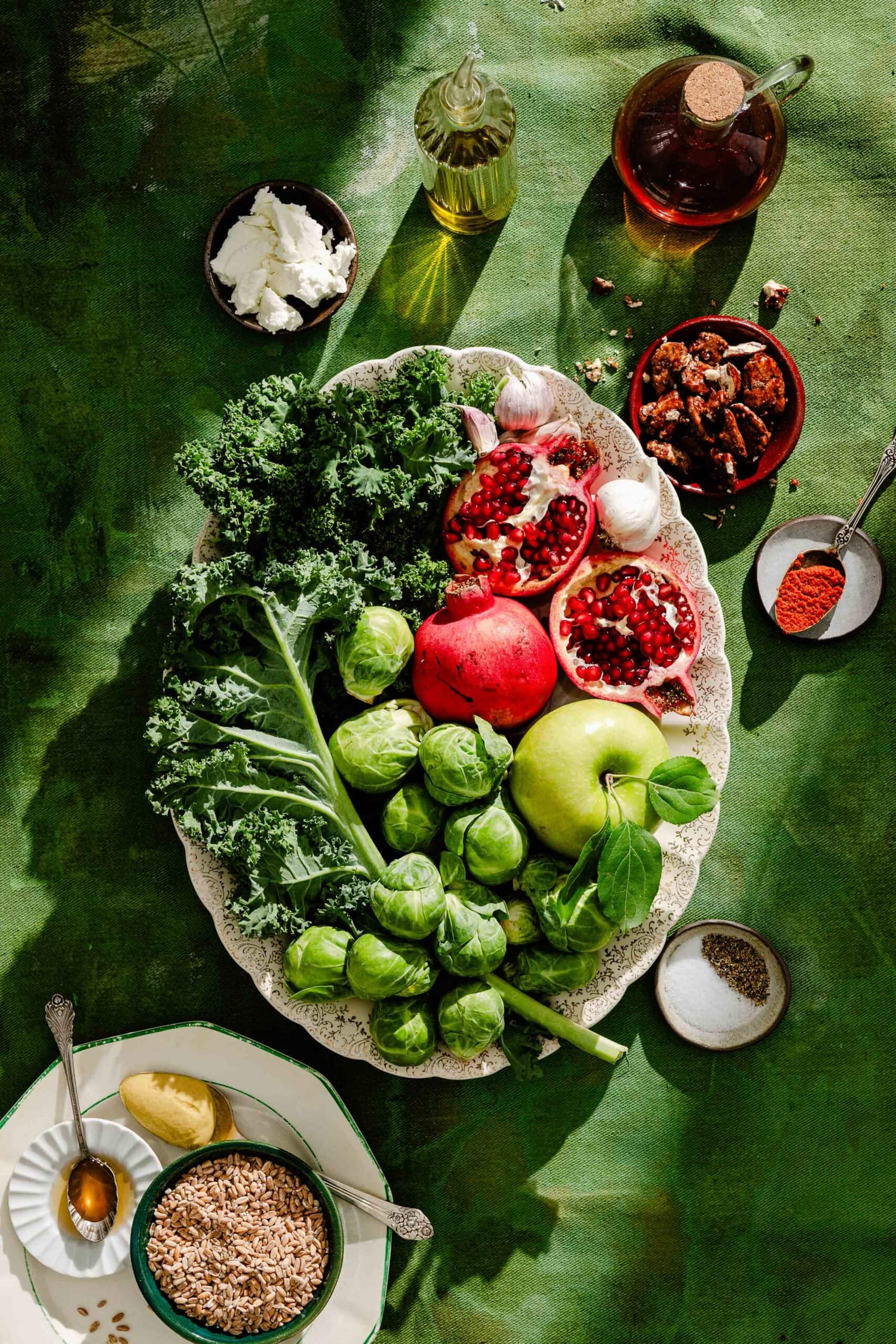 A large platter of kale, Brussels sprouts, pomegranate halves, and a green apple, surrounded by bowls of feta, spiced nuts, grains, oil, and seasonings on a green surface.