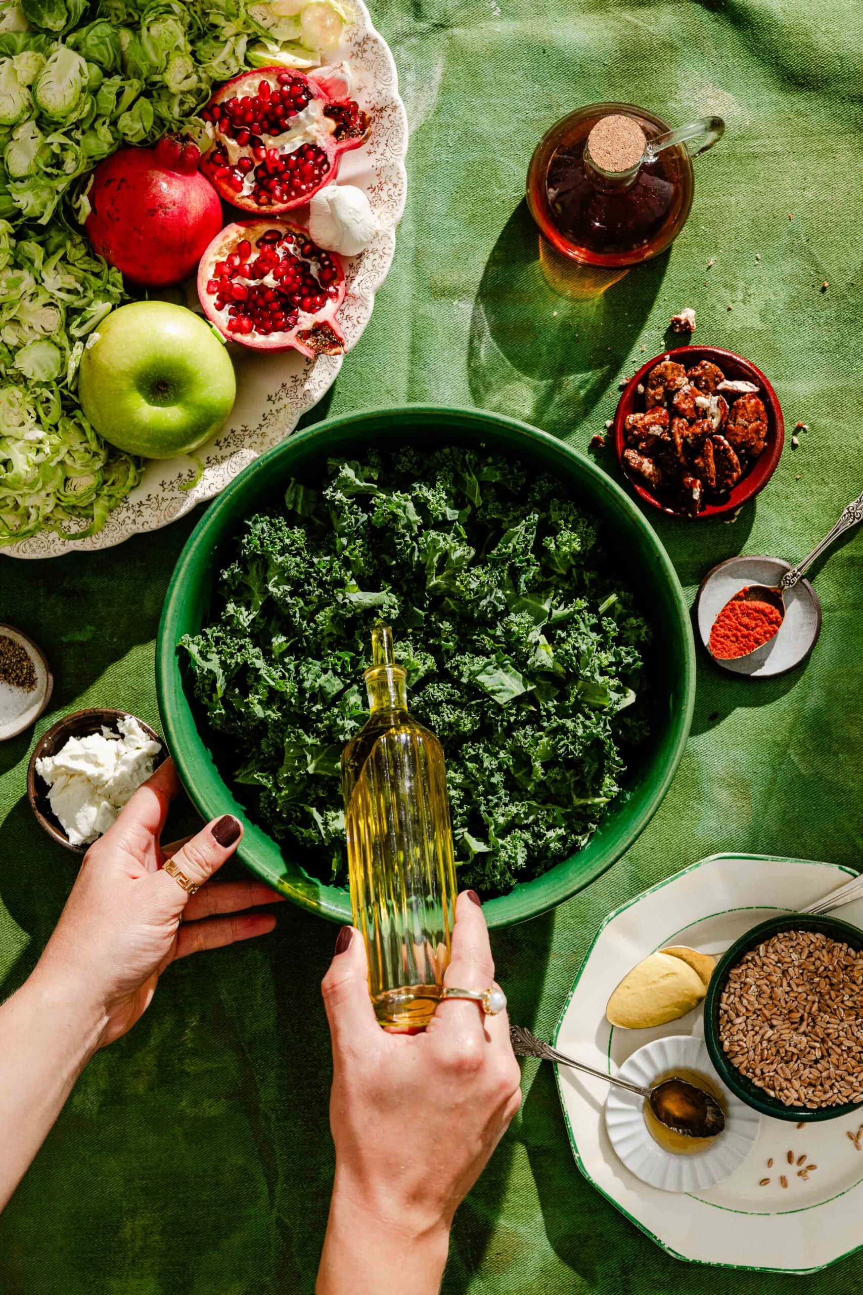 Hands pouring oil from a glass bottle into a large bowl of kale, surrounded by pomegranate, brussels sprouts, apple, nuts, cheese, and other ingredients on a green tablecloth.
